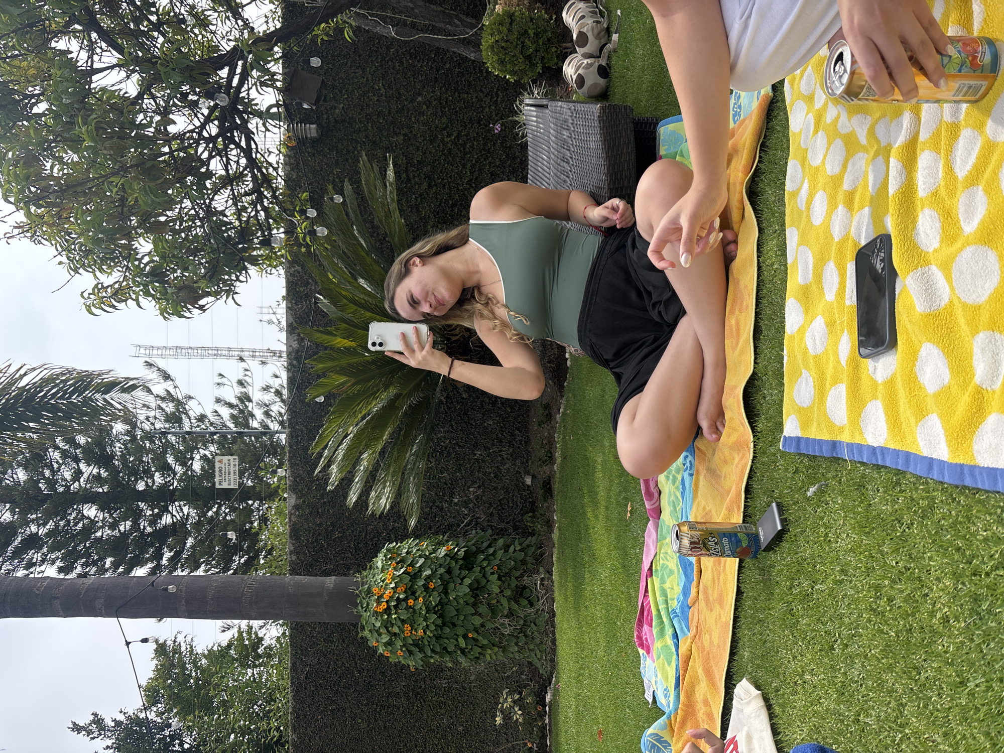 The scene is a residential backyard in Puebla, Mexico. Two individuals are present. One woman is holding a smartphone, presumably taking a photo or video. She is wearing a green tank top and black shorts. The second person is cropped in the image, wearing a white shirt. They are sitting on a yellow and white polka dot towel. Near the second person, there is an open can of beverage and a cell phone on the towel. There is green grass, a large palm tree, a hedge with orange flowers, and a power line sign overhead.