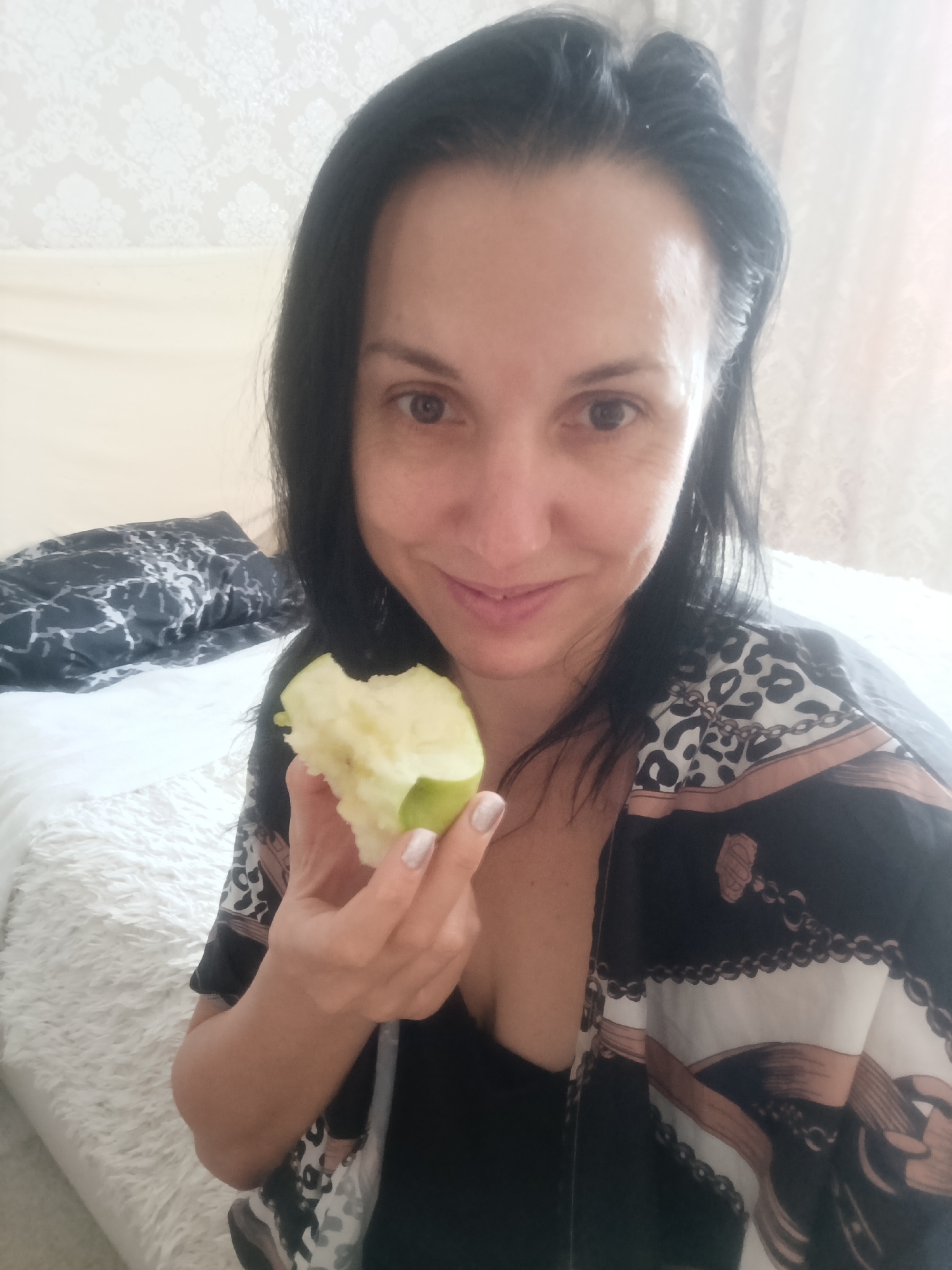 A woman with dark hair is holding a partially eaten green apple in her right hand. She is wearing a black top and a black and white patterned robe. She appears to be indoors, possibly in a bedroom, with a white, fluffy blanket visible behind her and a patterned duvet on a bed to her left. The background also shows a light-colored, textured wallpaper. The image does not contain information about the city of Penza, Russia.