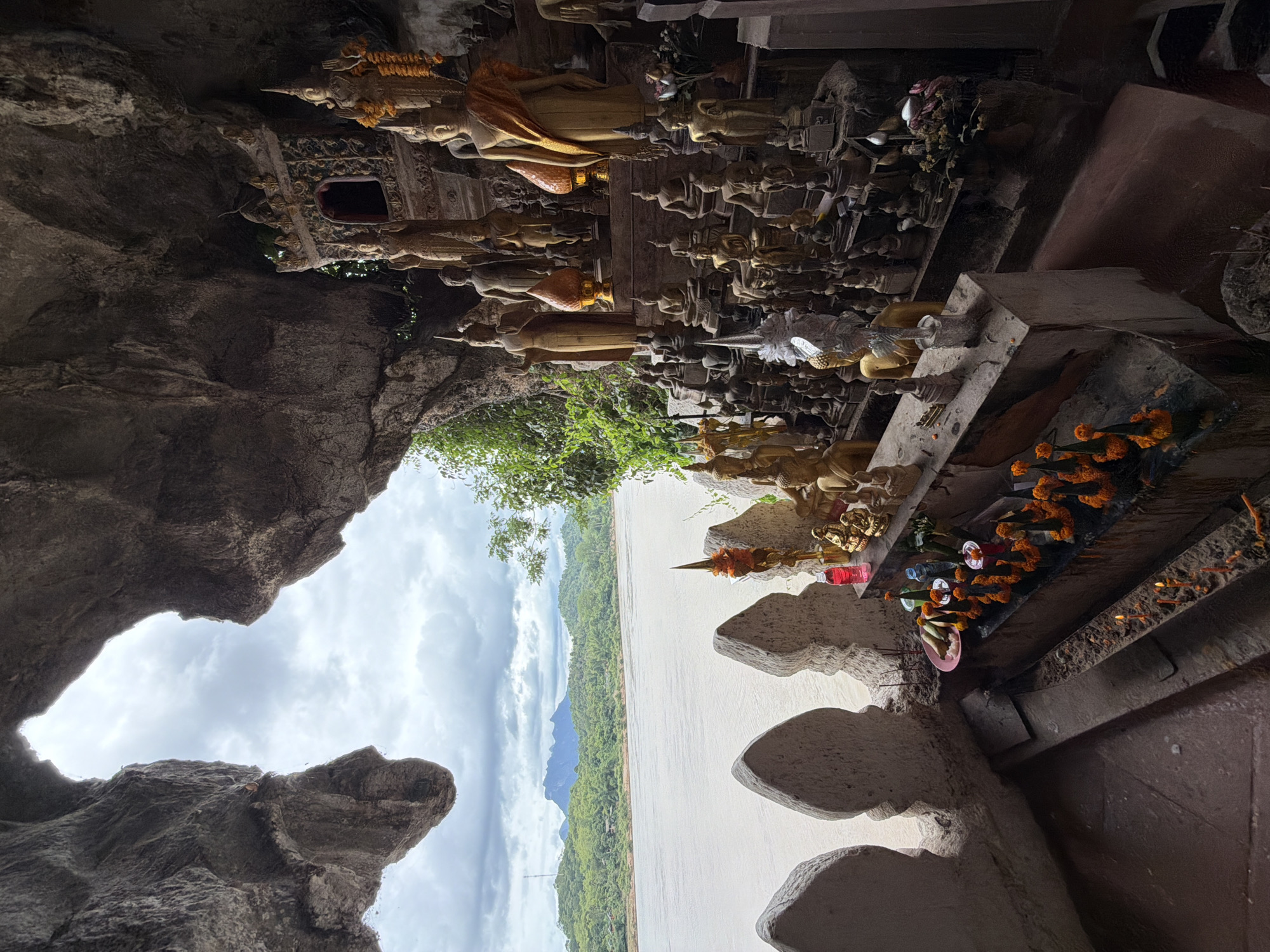The image shows the interior of a cave containing numerous Buddha statues and offerings. The cave opening frames a view of a wide river and lush, green mountains under a cloudy sky. In the foreground, various offerings are arranged on a stone ledge, including flowers, candles, and what appear to be small packets of food. There are multiple tiers of golden Buddha statues of varying sizes, some standing and some seated, adorned with orange sashes and garlands. In the middle ground, a stone wall with decorative cutouts overlooks the river. The environment is a natural cave setting with rough rock formations forming the ceiling and walls, contrasting with the carefully arranged religious artifacts. The scene is located within the city of Muangkeo, Laos, a place known for its numerous caves and temples.