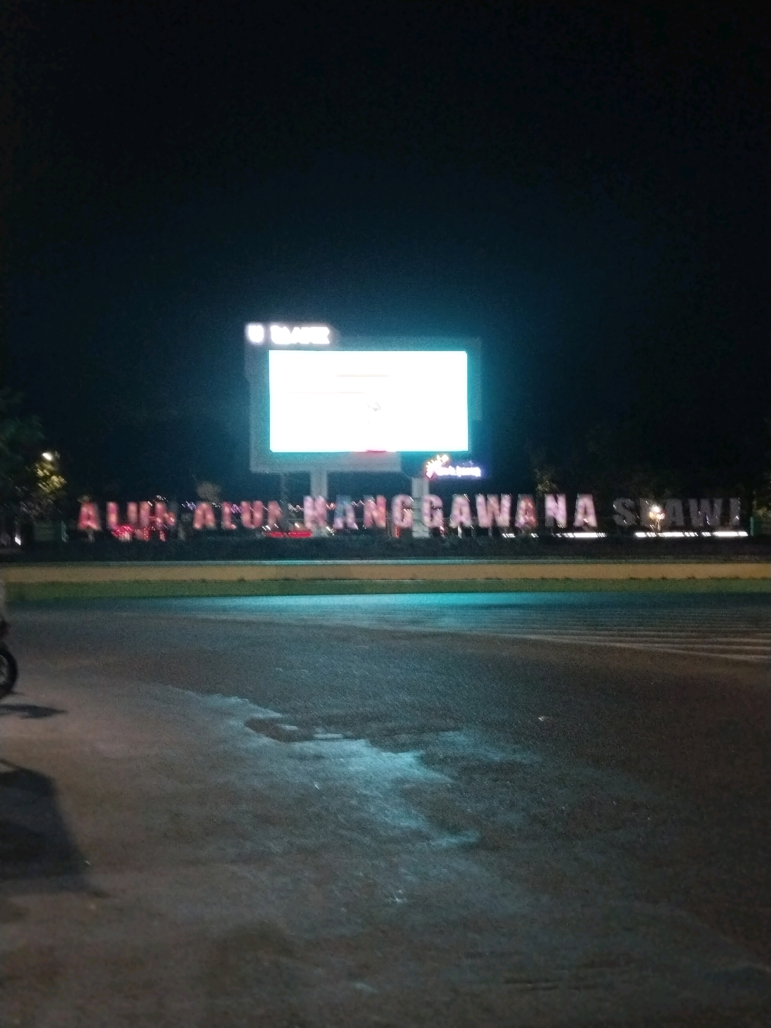 The image captures a nighttime scene at the Alun-Alun Sangawana in Slawi, Indonesia. In the background, a large, illuminated billboard displays white text against a dark background. Below the billboard, large, lit letters spell out "ALUN ALUN SANGGAWANA SLAWI". The foreground shows a wet asphalt road, with reflections of streetlights and possibly the illuminated letters on its surface. A shadow of a motorcycle is visible on the left side of the road. The overall environment suggests an outdoor public space at night, likely a town square or central gathering area.