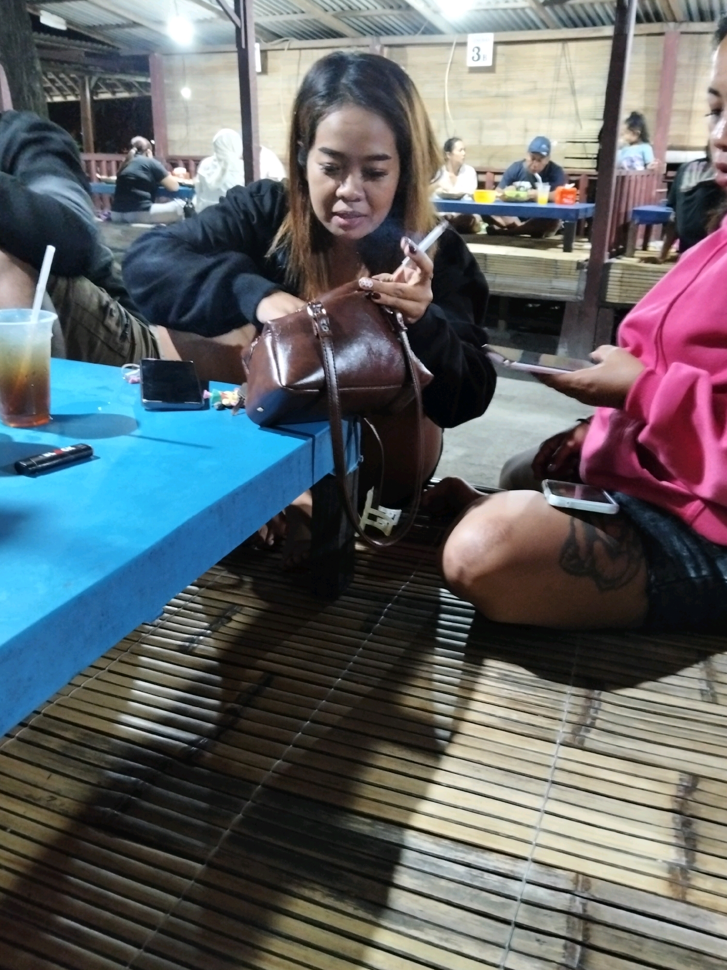 An outdoor, dimly lit open-air restaurant features a blue plastic table and a bamboo mat floor. Two women are seated at the table. The woman on the left is wearing a black top and is smoking a cigarette, holding a brown purse. A plastic cup with a drink and a straw is on the table, along with a phone and a remote. The woman on the right is wearing a pink top and is looking at her phone, which is placed on her thigh, revealing a tattoo on her leg. In the background, other patrons are seated at tables, and a sign with the number "3" is visible.