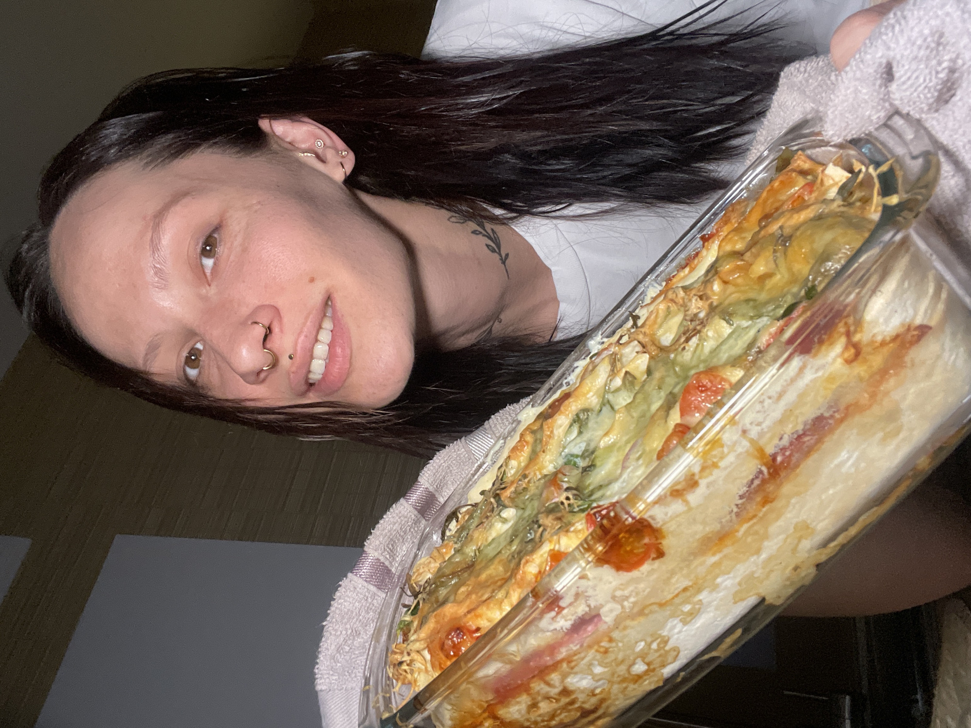 A woman with dark hair and multiple piercings, including in her nose and ears, holds a clear glass baking dish filled with what appears to be a layered lasagna-style dish. The food has visible layers of pasta, sauce, and vegetables. The dish is being held with a light-colored towel. The woman is wearing a white t-shirt and has a tattoo on her neck. The background is blurred, showing a wall with a wooden panel and a corner of a room. There is no information to suggest the picture was taken in Izhevsk, Russia.