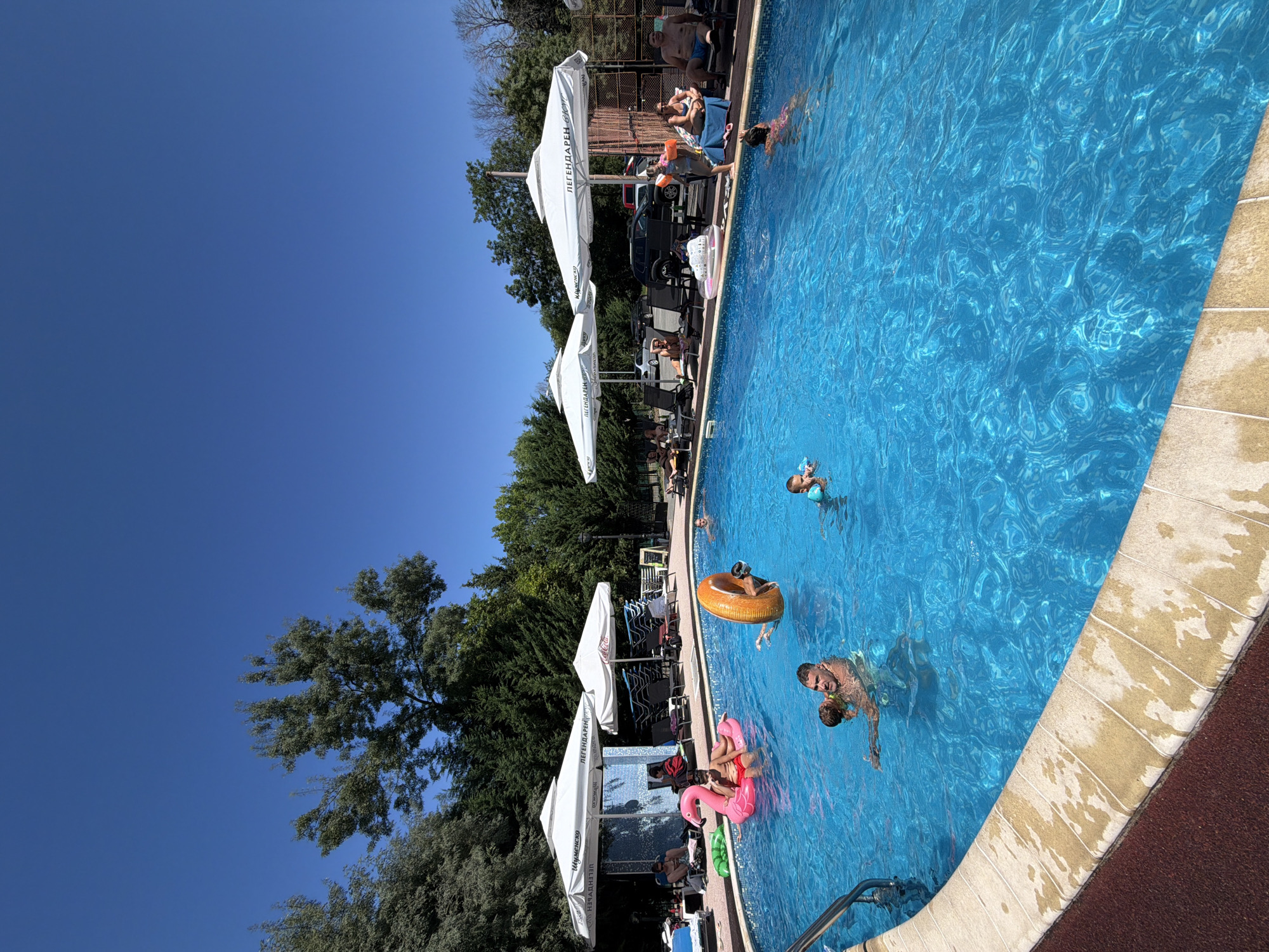 The image depicts a sun-drenched outdoor swimming pool scene at a location in Burgas, Bulgaria. The pool, filled with clear blue water, is the central focus. Several people are enjoying the water and the surrounding area. In the foreground, a man is holding a young child in the pool. To the left, a person is sitting on a pink flamingo float. In the middle of the pool, a person is holding onto an orange inflatable ring. Further back, other individuals are visible swimming or relaxing. The poolside is lined with lounge chairs and white umbrellas offering shade. Trees and foliage form a backdrop to the scene. The sky is a vibrant, clear blue. The location appears to be a recreational facility, possibly a hotel or resort, given the presence of umbrellas with branding, one of which reads "ЛЕГЕНДАРЕН ВКУС."
