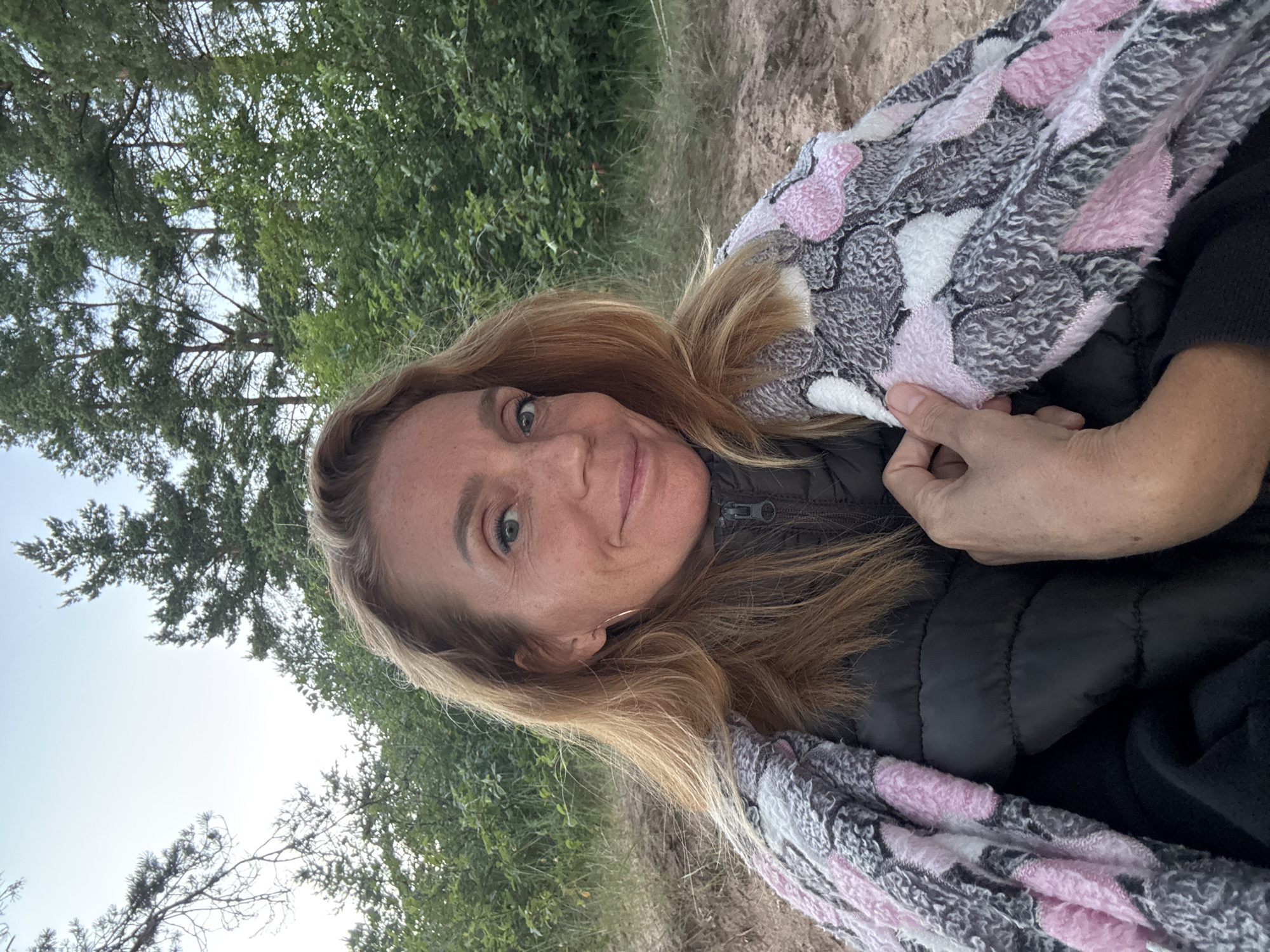This is a portrait-oriented, slightly over-the-shoulder selfie of a woman outdoors. She has long, blonde hair that falls around her face and shoulders. Her eyes are green, and she is smiling gently. She is wearing a black puffer jacket, and a patterned scarf with pink and gray elements is draped over her shoulder and chest. The background shows a natural setting with trees and what appears to be a sandy path or dune. The lighting suggests it is daytime, possibly late afternoon or early morning, given the soft light and muted colors. There is no visible text in the image. The woman's expression conveys a sense of contentment and peacefulness. The location cues of trees and sand suggest a natural outdoor environment, possibly a park or a coastal area.