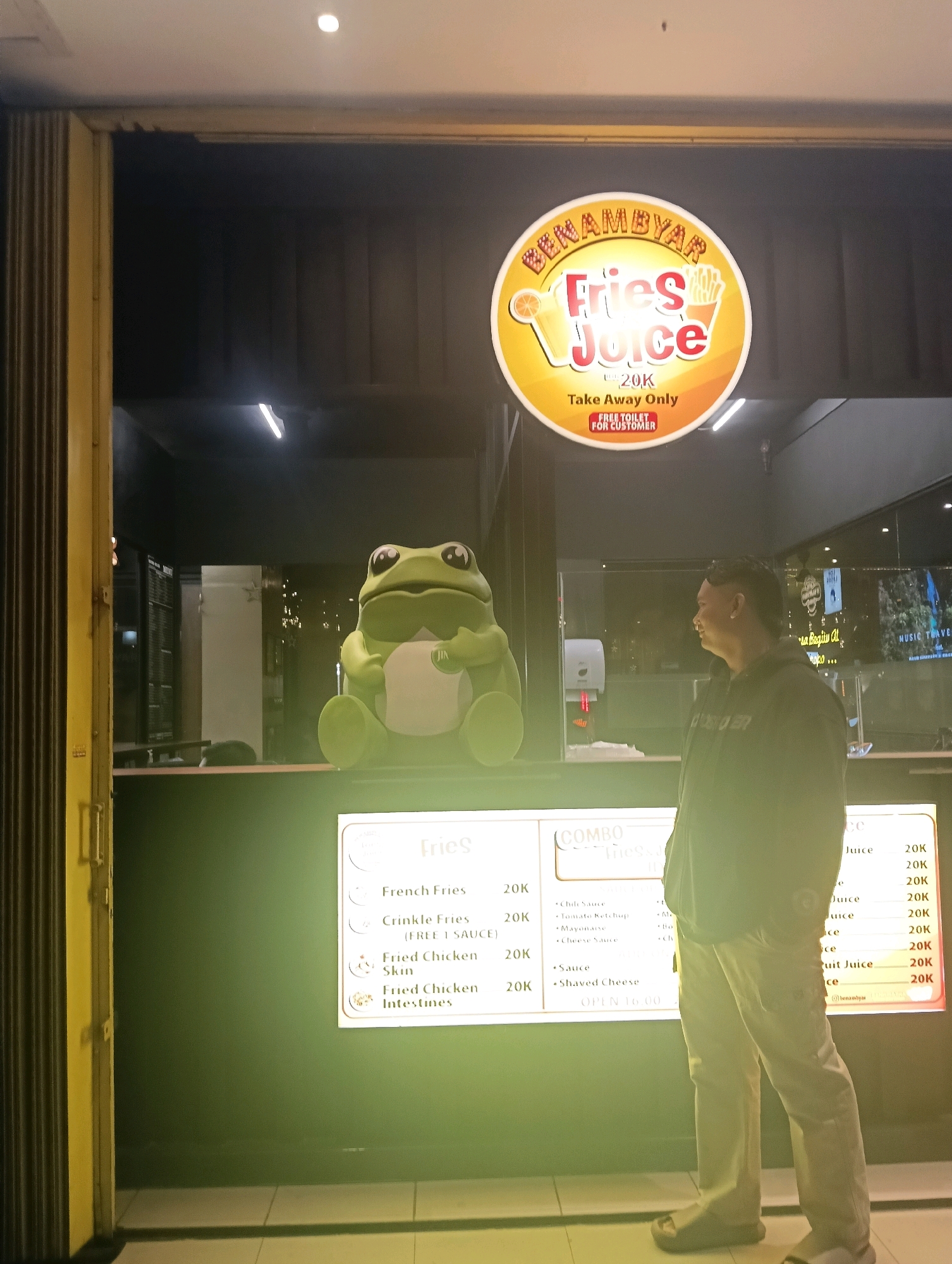An outdoor shot captures a man standing in front of a food stall in Malang, Indonesia, at night. The stall, named "BENAMBYAR Fries Juice," features a large, circular sign with a cartoon orange and fries graphic, advertising "Fries Juice" for 20K and "Take Away Only." A large, green, cartoon frog statue is placed on the counter. To the right, a menu board displays various fries and juice options with their prices. The man, wearing a dark hoodie and light-colored pants, is looking towards the menu board. The surrounding environment appears to be a street with dimmed lighting.