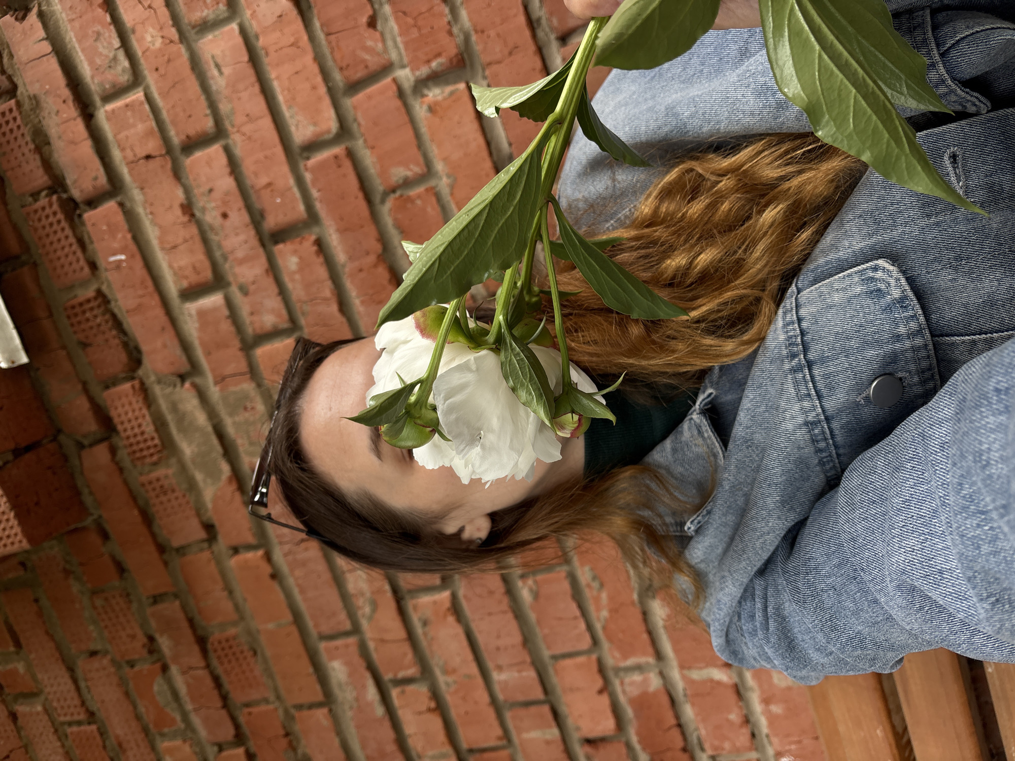 A young woman with auburn hair, wearing a denim jacket over a dark green top, is seated on a wooden bench in front of a red brick wall. She is holding a white peony flower, which partially obscures her face, with her right hand. Sunglasses are perched on her head. The background consists of a textured red brick wall. The scene appears to be outdoors, possibly in a courtyard or garden. The location is identified as Votkinsk, Russia.