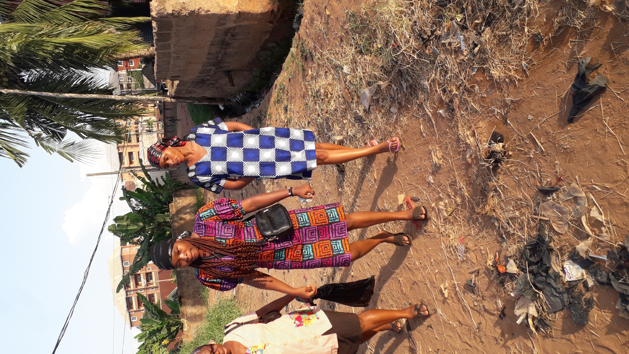 Three individuals are positioned on an earth-toned ground covered with dried vegetation and scattered trash. The person in the foreground is partially visible, wearing a light-colored, short-sleeved shirt and dark shorts, holding a black plastic bag. The second person is wearing a colorful dress with geometric patterns, carrying a black shoulder bag, and has long braided hair. The third person is wearing a blue and white checkered dress and a patterned headscarf. In the background, there are buildings, trees, and other urban elements. The location appears to be a roadside area within the city of Nkpor, Nigeria.