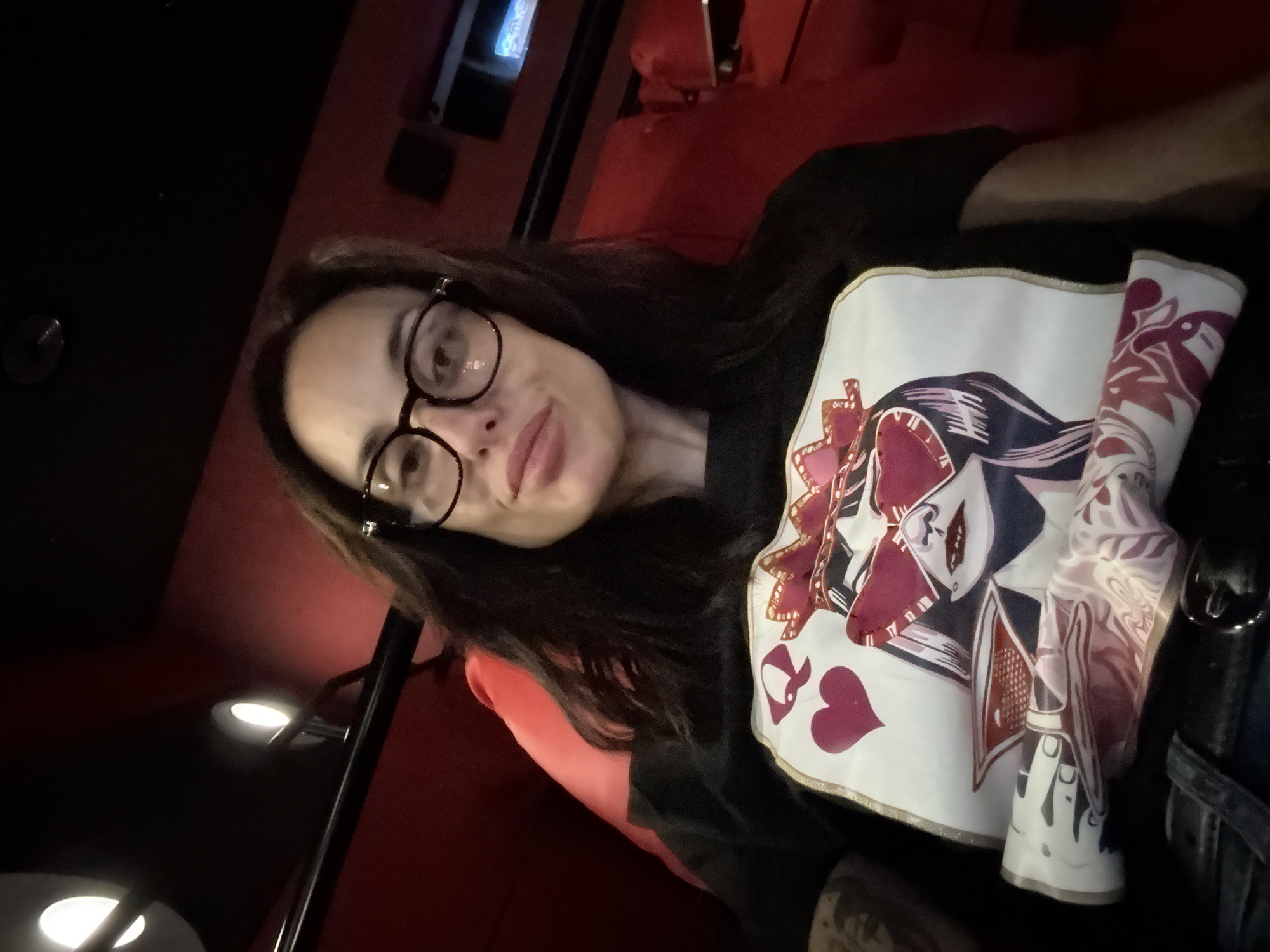A woman with long dark hair and glasses is posing for a selfie. She is wearing a black t-shirt with a white design featuring playing cards. The woman appears to be sitting in a red seat, possibly in a cinema or theater. There is a small television screen visible in the background. The lighting is dim. It is not possible to ascertain if this image was taken in Pleven, Bulgaria.