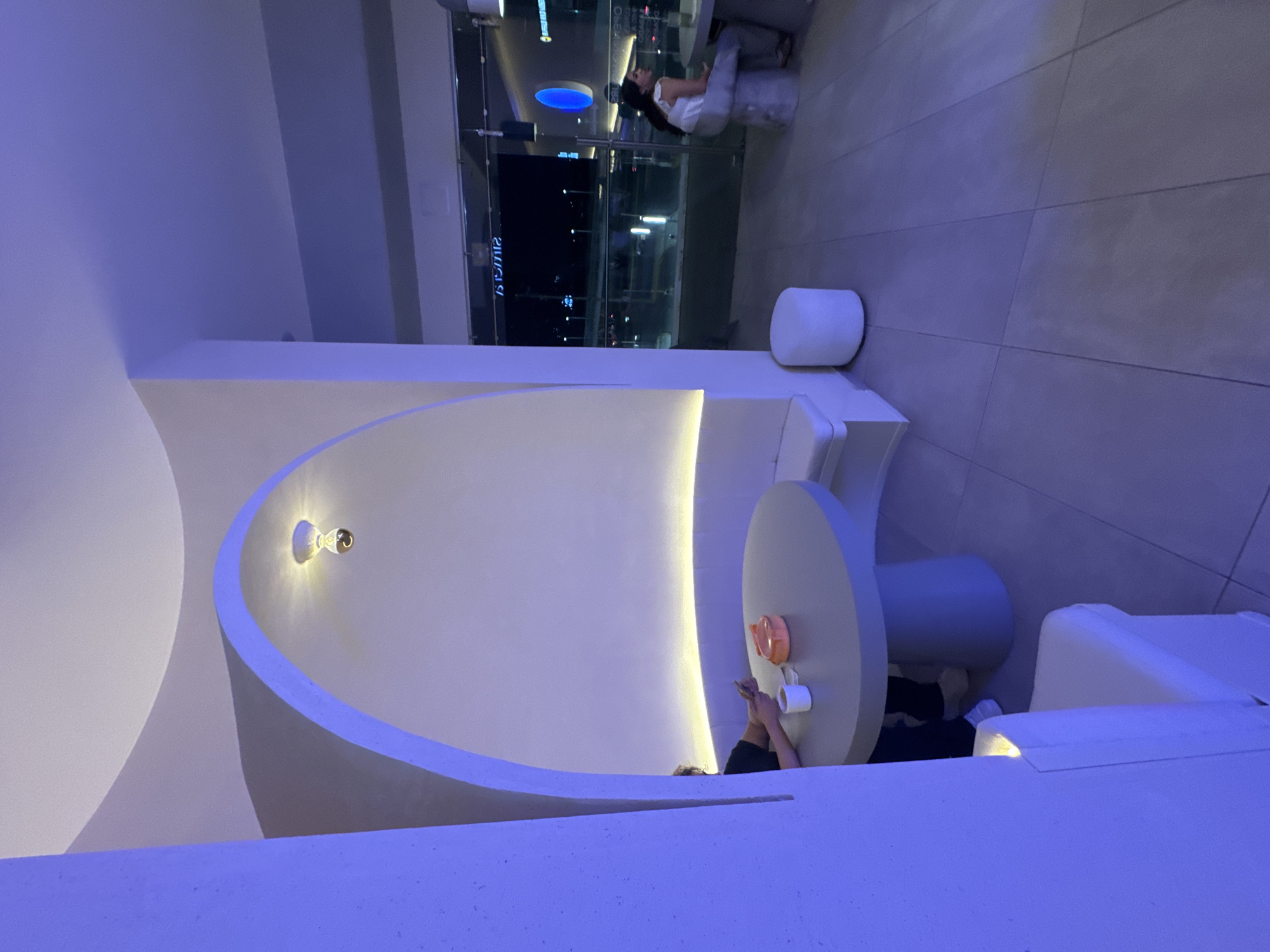 A modern, minimalist indoor setting is illuminated by soft, cool-toned lighting. A white, curved architectural element dominates the foreground, partially obscuring a circular table where a person sits using their phone. The table holds a plate and a cup. Adjacent to the table, a cushioned bench provides seating. In the background, a glass wall reveals an exterior urban view at night, complete with city lights and a person seated in a distant part of the establishment. The location could be a cafe or lounge, possibly in San Bernardino Tlaxcalancingo, Mexico.