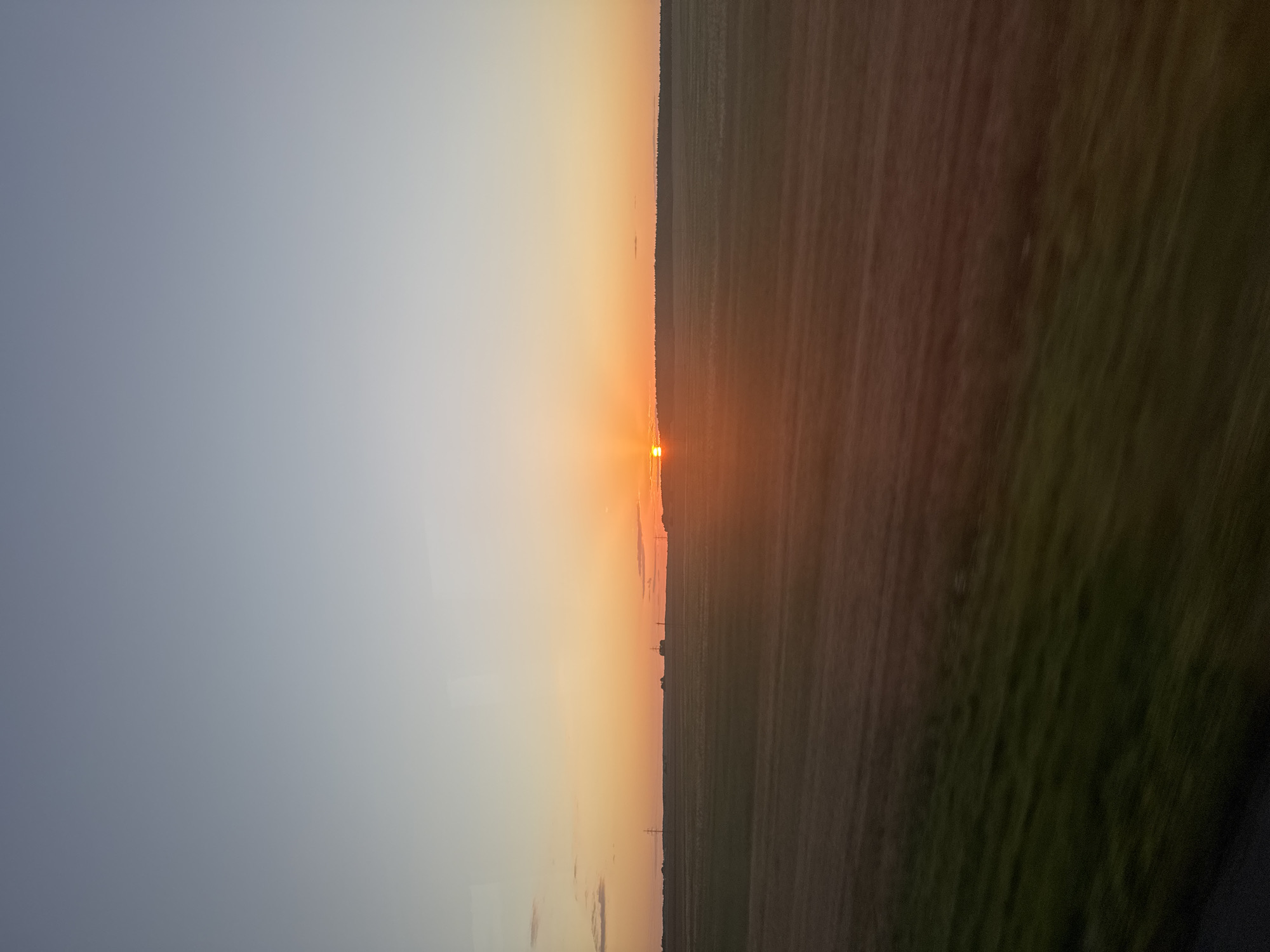 A wide shot captures a rural landscape at sunset. The sky transitions from a pale blue at the top to a warm orange and pink gradient near the horizon, with the sun setting just above a tree line. A vast, flat field stretches across the middle ground, appearing to have been recently harvested or tilled, with distinct rows visible. In the distance, faint silhouettes of power line towers can be observed. The foreground is a blur of green and brown, suggesting movement, possibly from a vehicle. The overall scene is serene and natural, with no visible people or structures other than the distant power lines. The location is identified as Сталовіцкі сельскі Савет, Belarus.