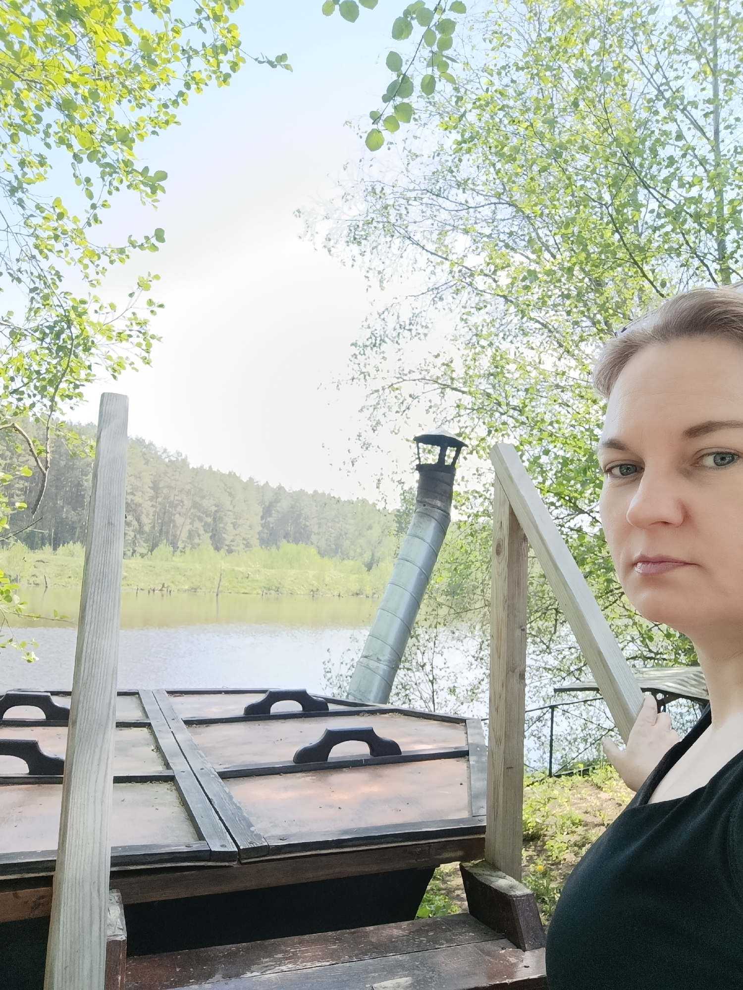 A woman stands in front of a wooden structure near a body of water in Prikamskii, Russia. The structure has a chimney and several rectangular lids with handles. Trees and a forest line the opposite shore of the water. The woman is looking towards the camera.