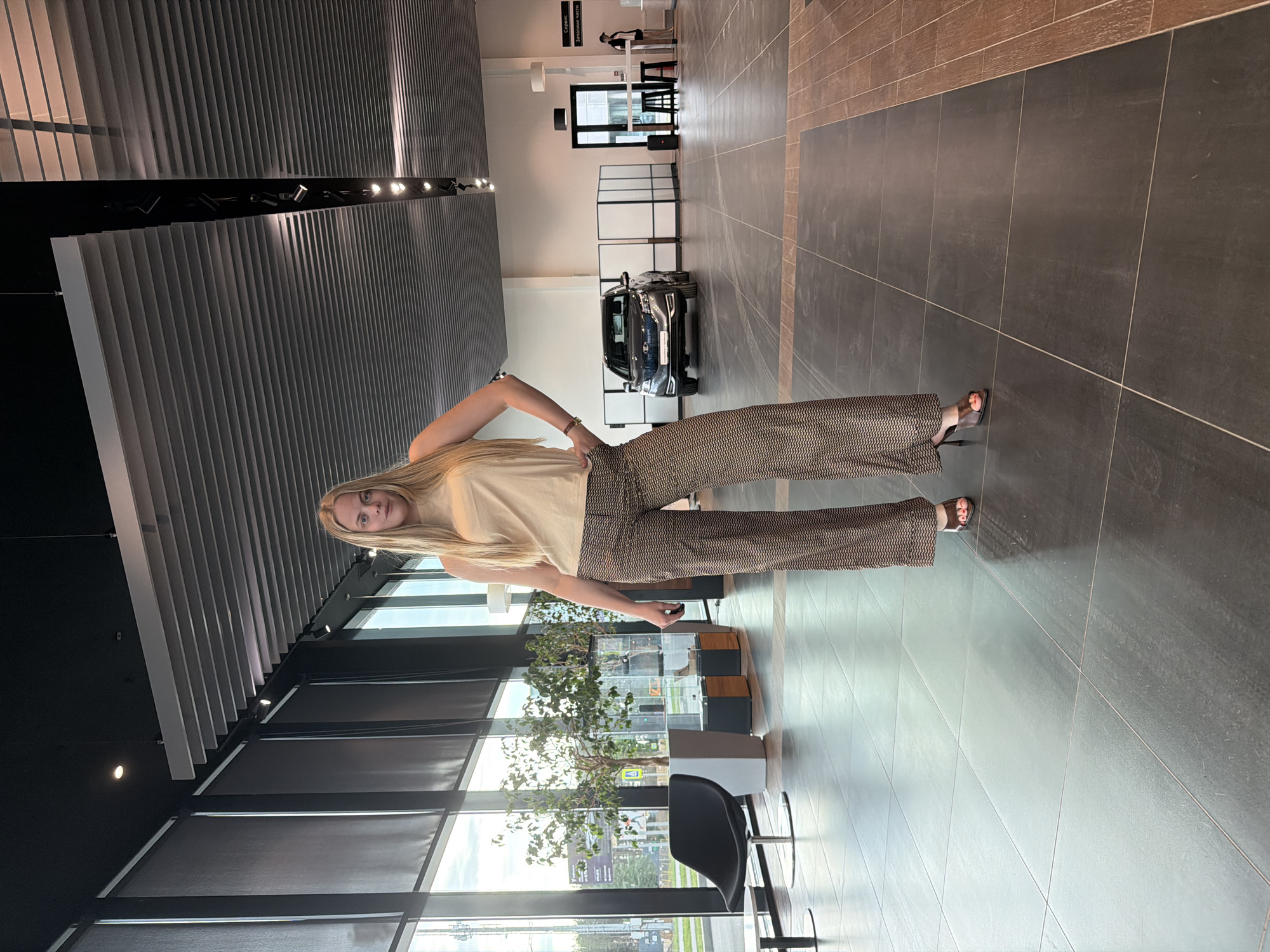 A full-body portrait of a young, fair-skinned woman with long blonde hair, wearing a light-colored top and patterned pants with dark sandals. She is standing in what appears to be an automotive showroom or dealership. The location is Izhevsk, Russia, as indicated by signage visible through the windows. In the background is a car, likely a vehicle for sale at the dealership. The showroom has large windows, a gray tiled floor, and modern lighting fixtures. Plants are located near the windows. A black chair can be seen near the plants.