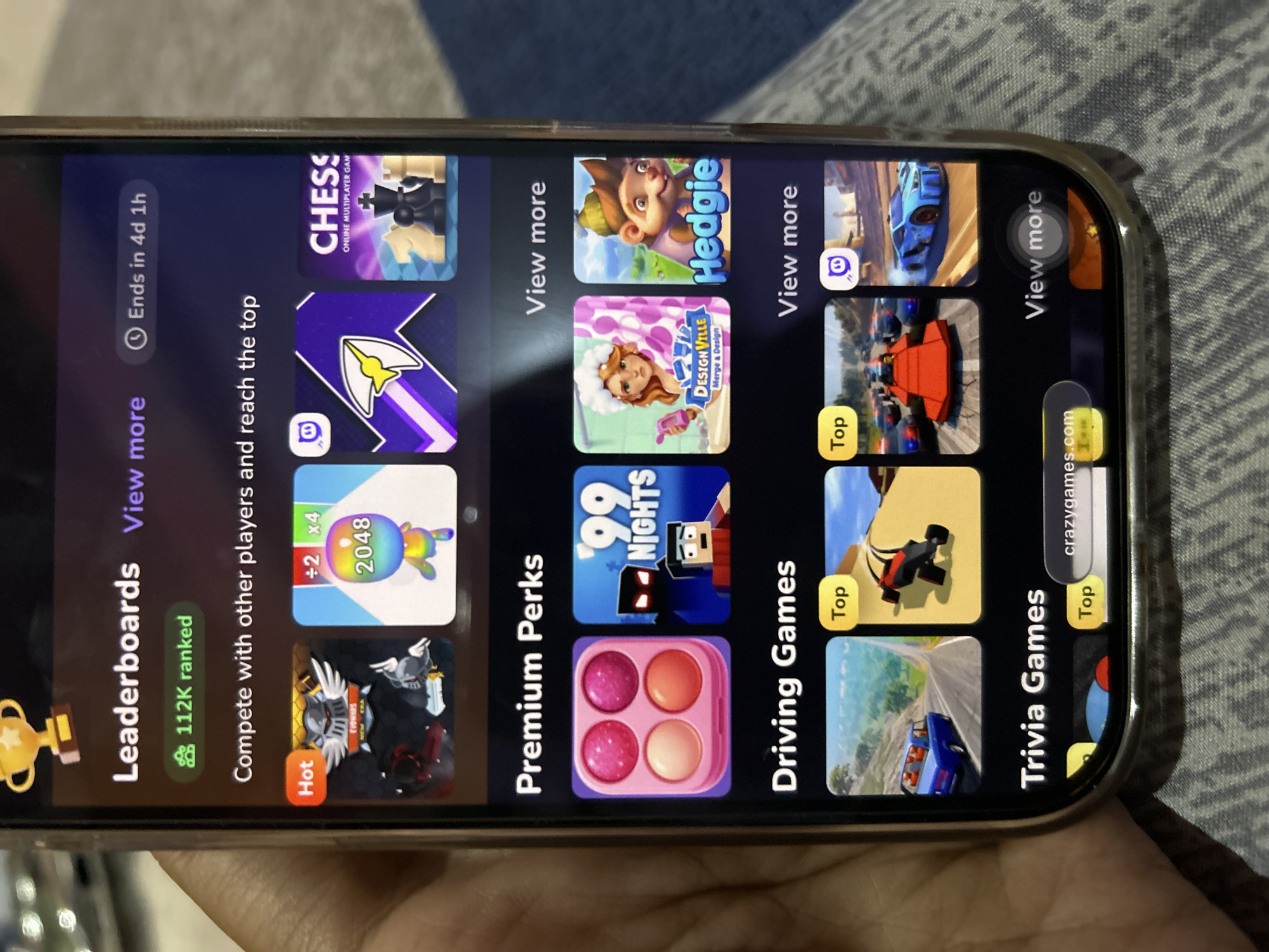 A close-up, eye-level shot captures a smartphone screen displaying a game application. The screen is dark purple and features various categories of games: "Leaderboards," "Premium Perks," "Driving Games," and "Trivia Games." Under "Leaderboards," there are several game icons, including one for "2048" and another for "Chess." "Premium Perks" shows icons for makeup products, a game titled "'99 Nights," and another called "Hedgie." "Driving Games" displays images of cars and racing. The "Trivia Games" section is partially visible. A hand holding the phone is visible at the bottom left of the frame. The environment appears to be indoors, with a patterned fabric in the background. There is no identifiable information linking this image to Depok, Indonesia.