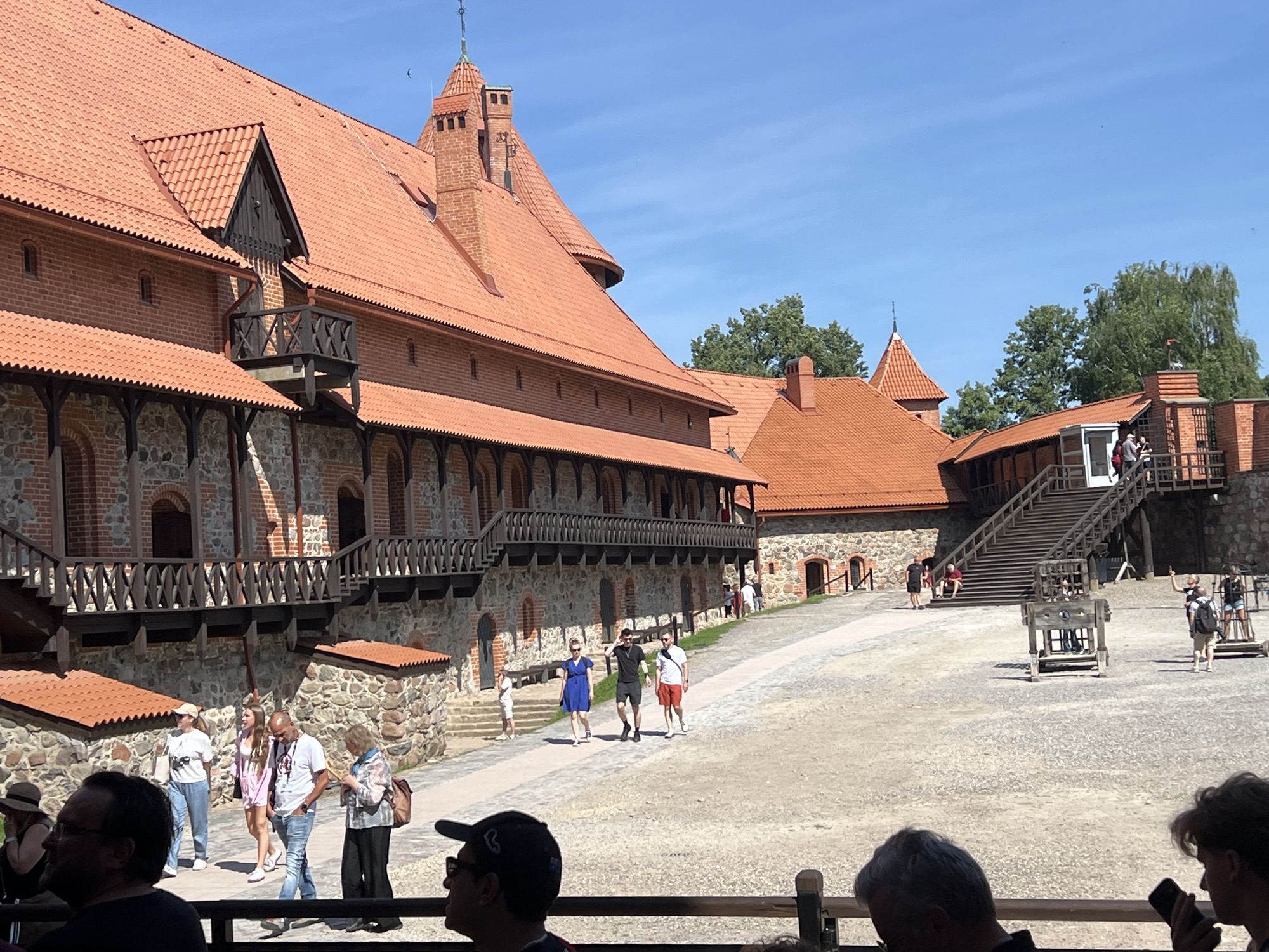 The image depicts the Trakai Island Castle in Trakai, Lithuania, on a sunny day. The castle, constructed of red brick with distinctive red-tiled roofs and wooden balconies, dominates the left side of the frame. Multiple tourists are visible walking around the castle grounds. In the foreground, several people are seated or standing, appearing to observe the scene. In the middle ground, a group of tourists walks along a path leading towards the castle. Further back, a set of wooden stairs ascends to a higher level of the castle. A historical pillory-like structure is present in the courtyard. The ground is covered in gravel and sand, and trees are visible in the background. The sky is clear and blue.