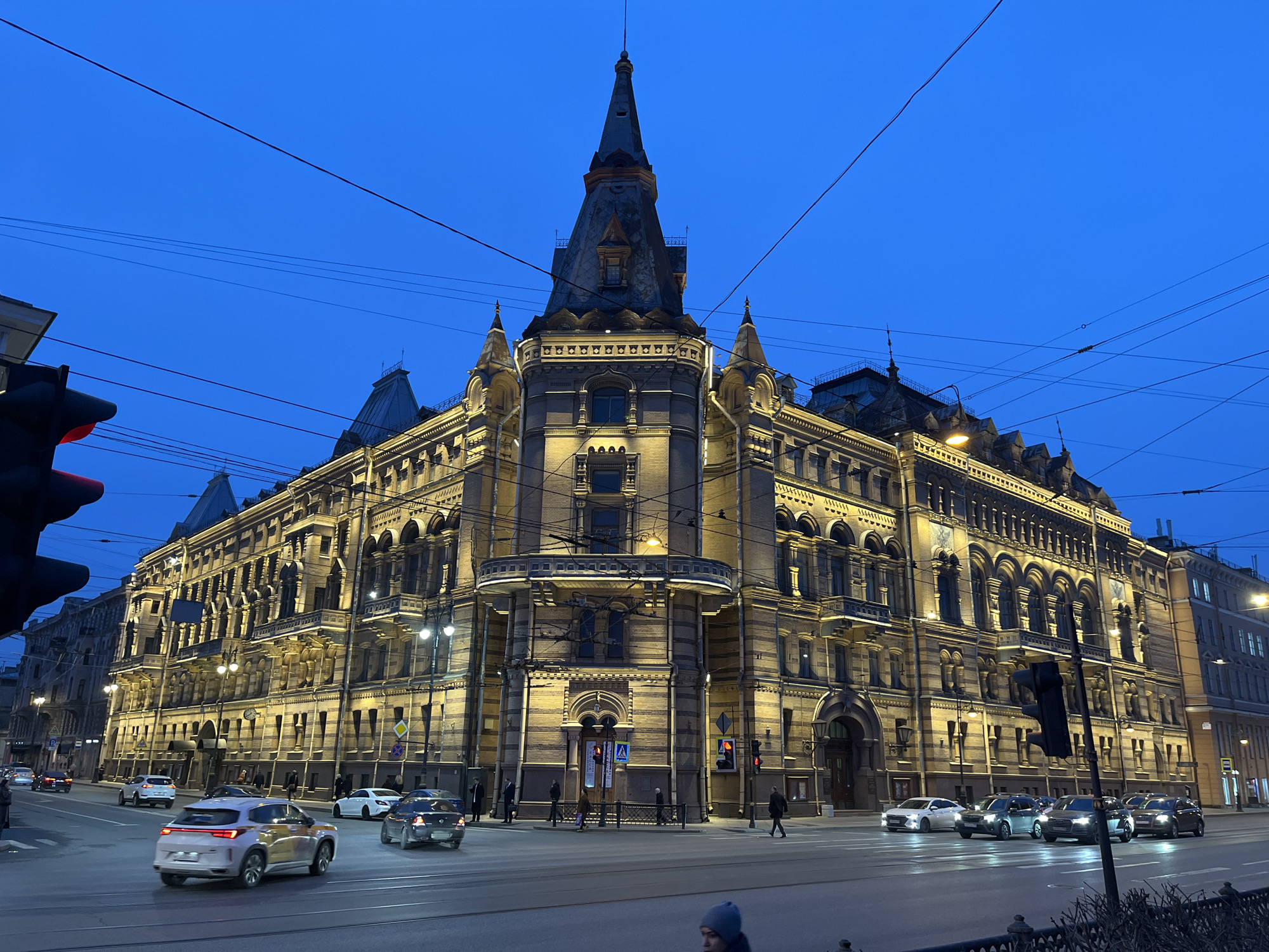 This image displays an urban street scene at night, focused on the Singer House (Dom Knigi) at Nevsky Prospekt 28, Saint Petersburg, Russia. The multi-story building, an architectural landmark featuring an ornate facade, a prominent corner turret, and a distinctive pointed spire, is extensively illuminated with warm yellow artificial lighting, contrasting with the deep blue, clear sky.
Nevsky Prospekt, a wide, multi-lane asphalt street, occupies the foreground. Several vehicles are in motion, including a light-colored SUV in the left-center lane and multiple other sedans and SUVs across various lanes. Overhead electrical wires, likely for trolleybuses or trams, are visible crisscrossing the upper portion of the frame. Multiple traffic light poles are present, with visible red signals.
Pedestrians are observed on the sidewalks adjacent to the building and at a marked pedestrian crossing. A partially cropped individual wearing a dark beanie is visible in the lower foreground. Decorative black metal fencing lines sections of the sidewalk. Streetlights cast additional illumination onto the roadway and surrounding areas.
