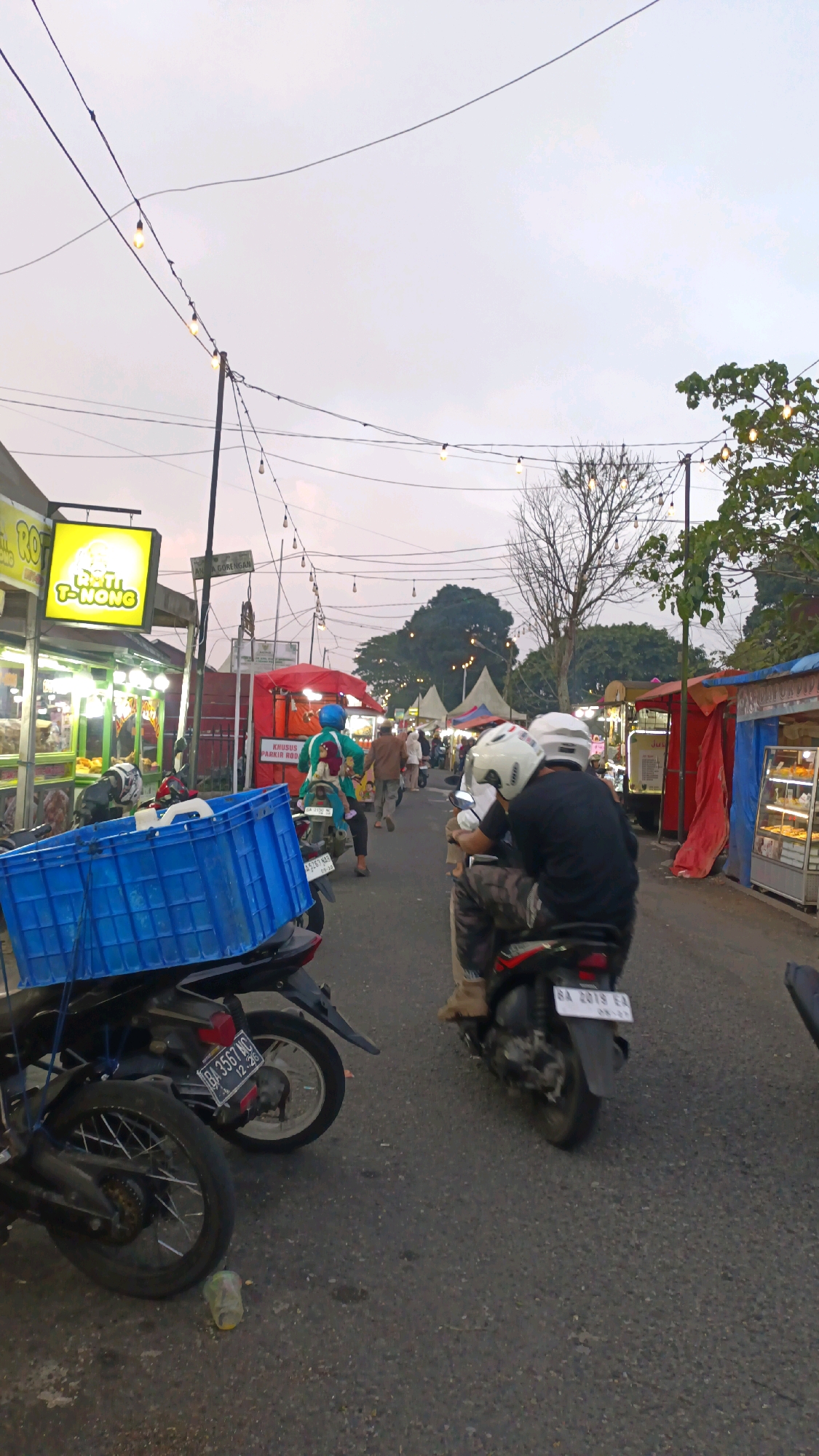 This image captures a street scene in Padang Panjang, Indonesia, likely during a twilight market or event. The foreground features several motorcycles parked or moving, one with a large blue crate attached to the back. Two individuals on a motorcycle, wearing helmets, are visible in the center-right of the frame, with the passenger leaning forward. Behind them, the street is lined with various stalls and shops, indicated by awnings, banners, and displayed goods. String lights are strung overhead, illuminating the street. People are seen walking and interacting at the stalls, suggesting a lively atmosphere. The background shows trees and buildings, with a hazy sky overhead. The overall impression is a bustling urban setting at dusk, with commerce and community activity taking place.