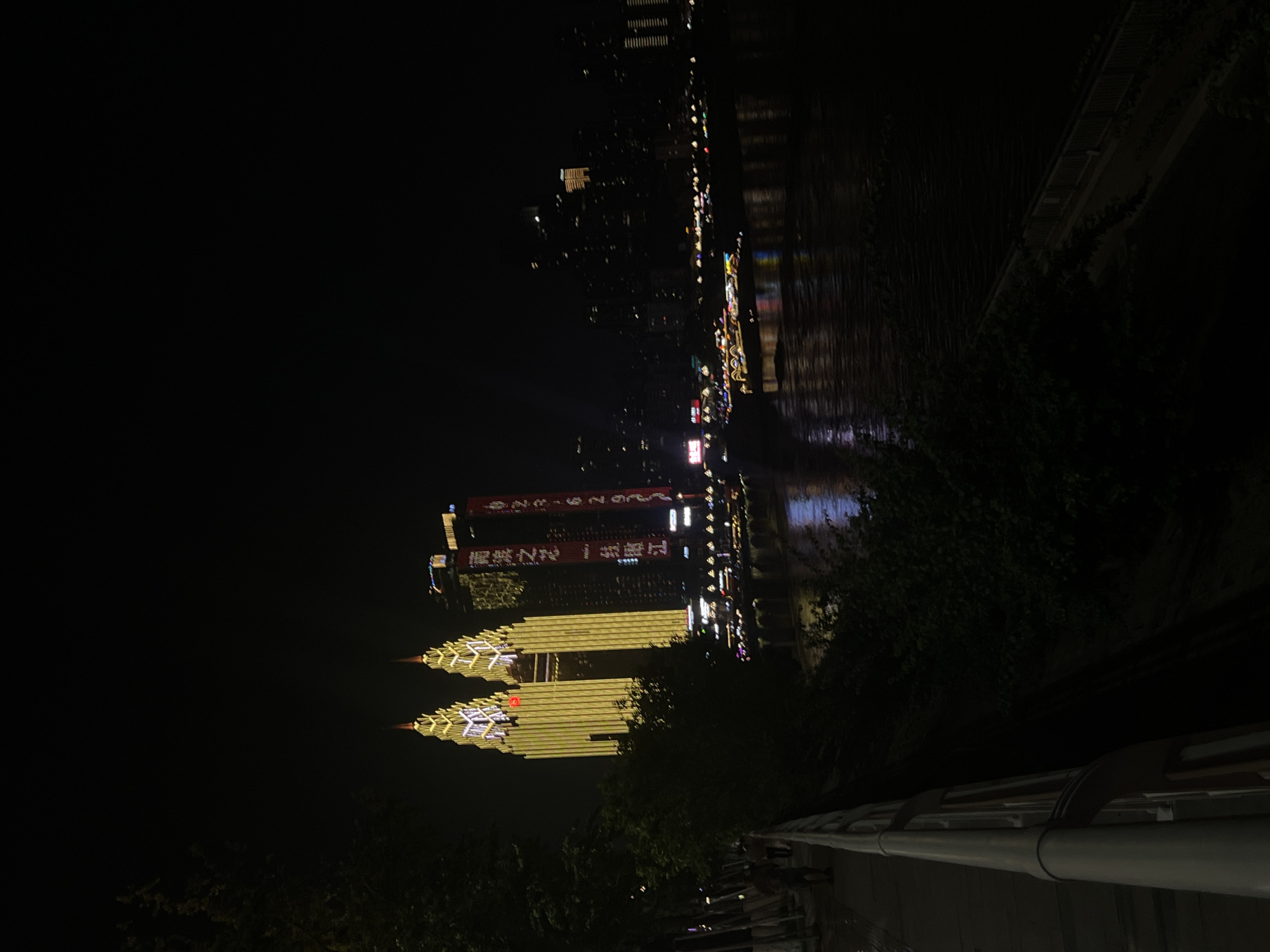 At night in Nan'an District, China, a wide river reflects the illuminated cityscape. Two prominent twin skyscrapers, adorned with bright yellow lights that outline their distinctive pointed tops, dominate the scene. A smaller red circular emblem is visible on the left skyscraper. To the right of these, other buildings rise, one with vertical red banners displaying white text. Boats with colorful lights are visible on the river, and their reflections shimmer on the water's surface. In the foreground, a dark, tree-lined embankment slopes down to the river. A walkway with a light-colored railing runs along the embankment, and a few pedestrians are seen walking on it. The overall impression is of a vibrant, modern city at night.