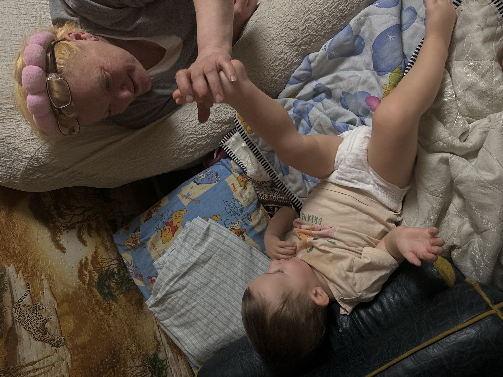 A high-angle shot depicts an adult holding the hand of a baby, with both figures lying down. The adult, an older woman with blonde hair and glasses, wears a pink padded headband and a gray top, her gaze fixed on the baby. The baby, dressed in a beige shirt and diaper, lies in a supine position with arms raised. They rest upon a couch, pillows, and blankets with varying patterns, including Winnie-the-Pooh and an animal print. The location is in the city of Unknown, Unknown.