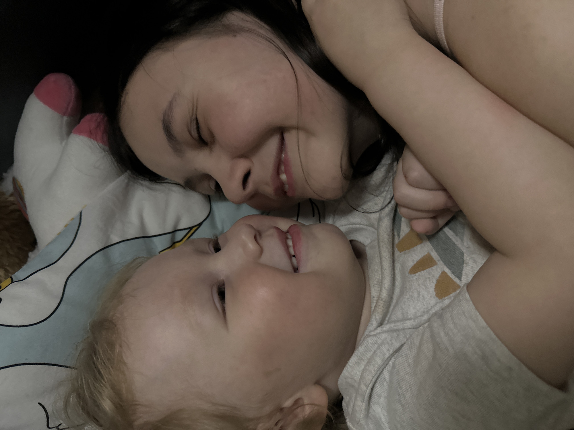 Two individuals are lying on a bed covered with a light blue and white patterned sheet. One is a female with dark hair and a light pink top. The other is a child with light blond hair and a grey and white shirt. Both are smiling and facing each other. A stuffed animal with pink and white socks is near their heads. The location is not indicated in the image. The location is unknown without additional information.