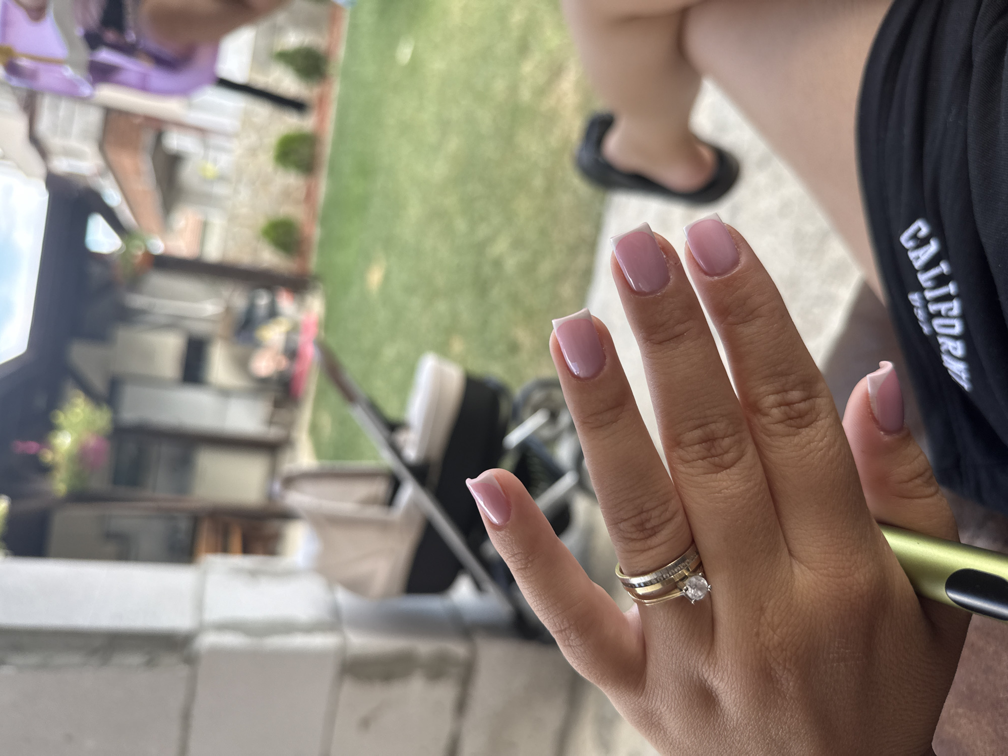 An outdoor setting, likely a backyard in Sandanski, Bulgaria, is depicted. The foreground focuses on a person's hand with freshly manicured pink nails and rings, including a diamond engagement ring. To the right, another person's legs are visible, wearing black shorts with white lettering. In the background, a stroller is present on a grassy area, suggesting a family or child-friendly environment. The overall scene conveys a relaxed, casual atmosphere, possibly during daytime given the bright lighting.