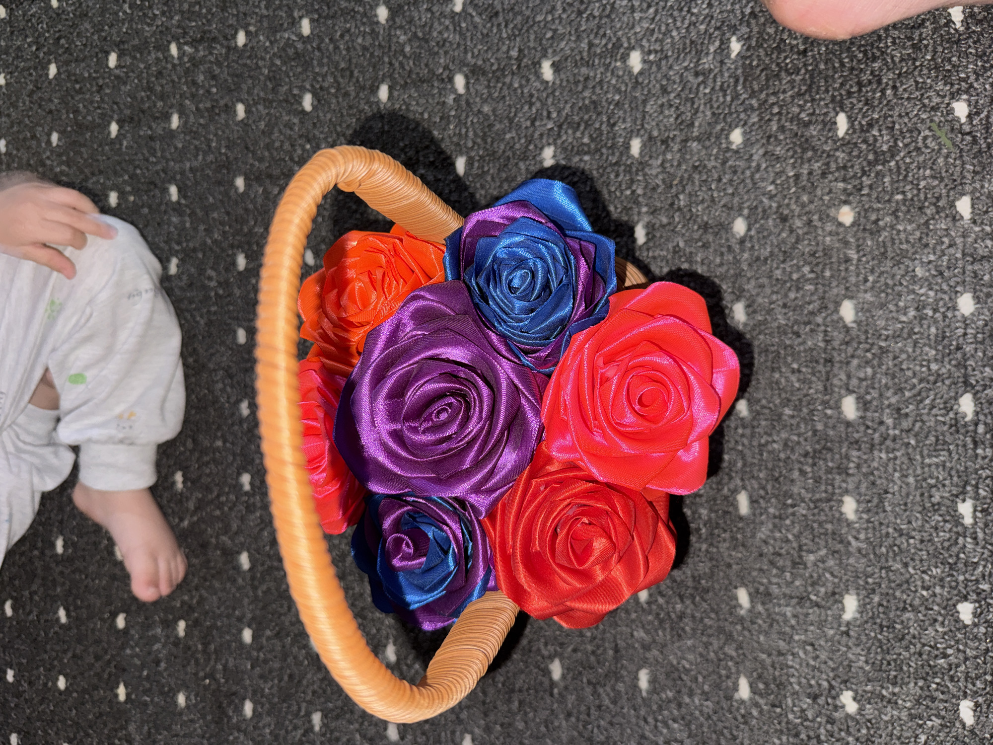 A close-up overhead shot shows a woven orange basket filled with fabric roses in various bright colors: red, pink, purple, blue, and orange. The basket is placed on a dark gray carpet with a scattered white dot pattern. In the blurred background, a person's legs and feet, clad in white pants, are visible. There is no specific indication of the location being Chardara, Uzbekistan.