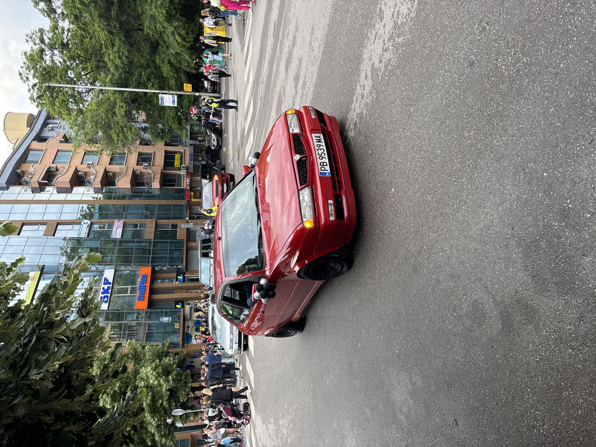The image shows a red Lancia Dedra Integrale car parked on a wet city street in Plovdiv, Bulgaria. The car has the license plate PB 9539 MA. People are gathered on the sidewalks beside the building, suggesting a parade or celebration is taking place. Buildings with glass fronts displaying names like Junker, SKF, and Credit Plus are visible in the background. Trees are also present on both sides of the street. A pedestrian crossing is marked on the street.