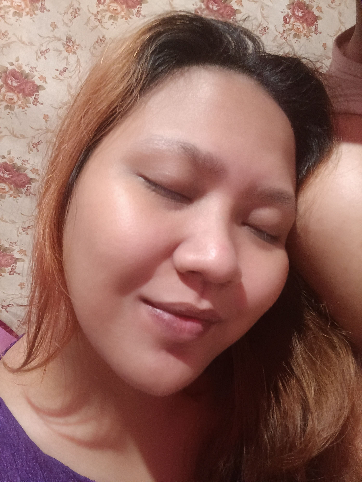 The image is a close-up selfie of a young woman lying down, with her eyes closed. She has light brown hair with some lighter streaks, and her skin appears smooth and slightly flushed. She is wearing a purple top. The background is a wall with a floral wallpaper pattern in muted tones of pink, cream, and green. To the right of the woman's head, another person's shoulder and arm are visible, suggesting they are lying close together or hugging. The lighting is soft and warm, indicating it might be daytime indoors or evening. The overall mood is relaxed and intimate. There is no visible text in the image. The location context is Depok, Indonesia.