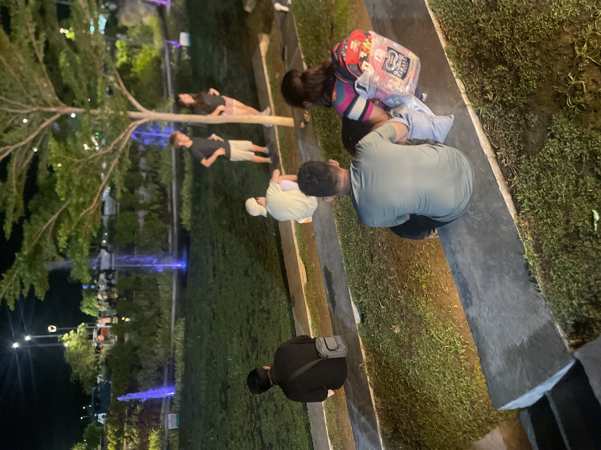 The image shows a group of people in a park at night. The location appears to be Unknown, Unknown, given the information provided. Several people are visible: one squatting with a gray bag over his shoulder, another kneeling with a backpack, two sitting or lying near a tree, and one sitting or lying farther down the line. A fountain is visible in the background with blue lights. The people appear to be relaxing in the park environment.