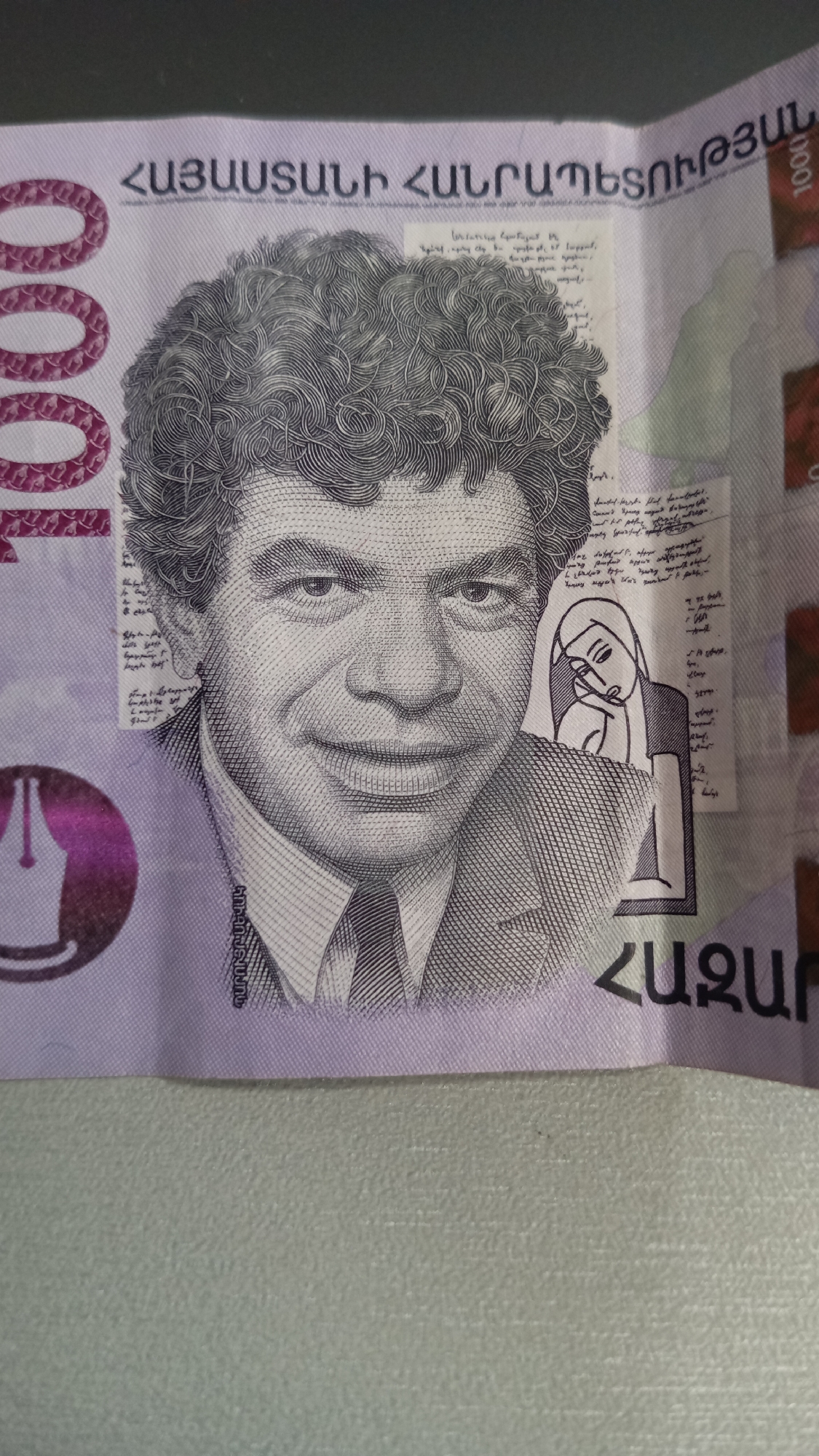 This image is a close-up of an Armenian banknote. On the banknote, a portrait of a man with curly hair is featured prominently. He is wearing a suit and tie. To the left of the portrait, the numeral "1000" is repeated vertically in a large font. Behind the portrait, there are lines of text that appear to be written in Armenian script. There is also a small illustration of a pen nib in a circle, colored purple. To the right of the portrait, there is a faint drawing of a woman with her head resting on her hand, also superimposed over the text. The banknote has a predominantly purple and white color scheme. The location in the city of Ashtarak, Armenia is not depicted in this image.