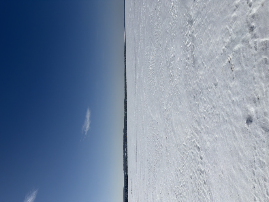 The image displays an expansive winter landscape, specifically a vast, snow-covered frozen body of water located near Votkinsk, Russia. The upper two-thirds of the frame is dominated by a clear, deep blue sky, featuring two distinct, wispy white clouds: one elongated cloud is situated in the upper-left quadrant, and a smaller, more compact cloud is visible slightly above the distant horizon line in the middle-right.
A sharp, dark horizon line, consistent with a distant treeline or shoreline with sparse structures, stretches horizontally across the entire width of the image, separating the sky from the snow-covered foreground. The lower third of the frame reveals a wide, relatively flat expanse of snow, exhibiting subtle undulations, ripples, and textured patterns across its surface, indicative of wind or foot traffic.
In the far distance, on the snow-covered surface near the right-hand side of the horizon line, three small, dark, indistinct figures are discernible. These figures appear to be stationary objects or individuals, potentially engaged in activities such as ice-fishing. The scene is brightly illuminated by direct sunlight, characteristic of a clear winter day, with no prominent shadows cast across the snow.
