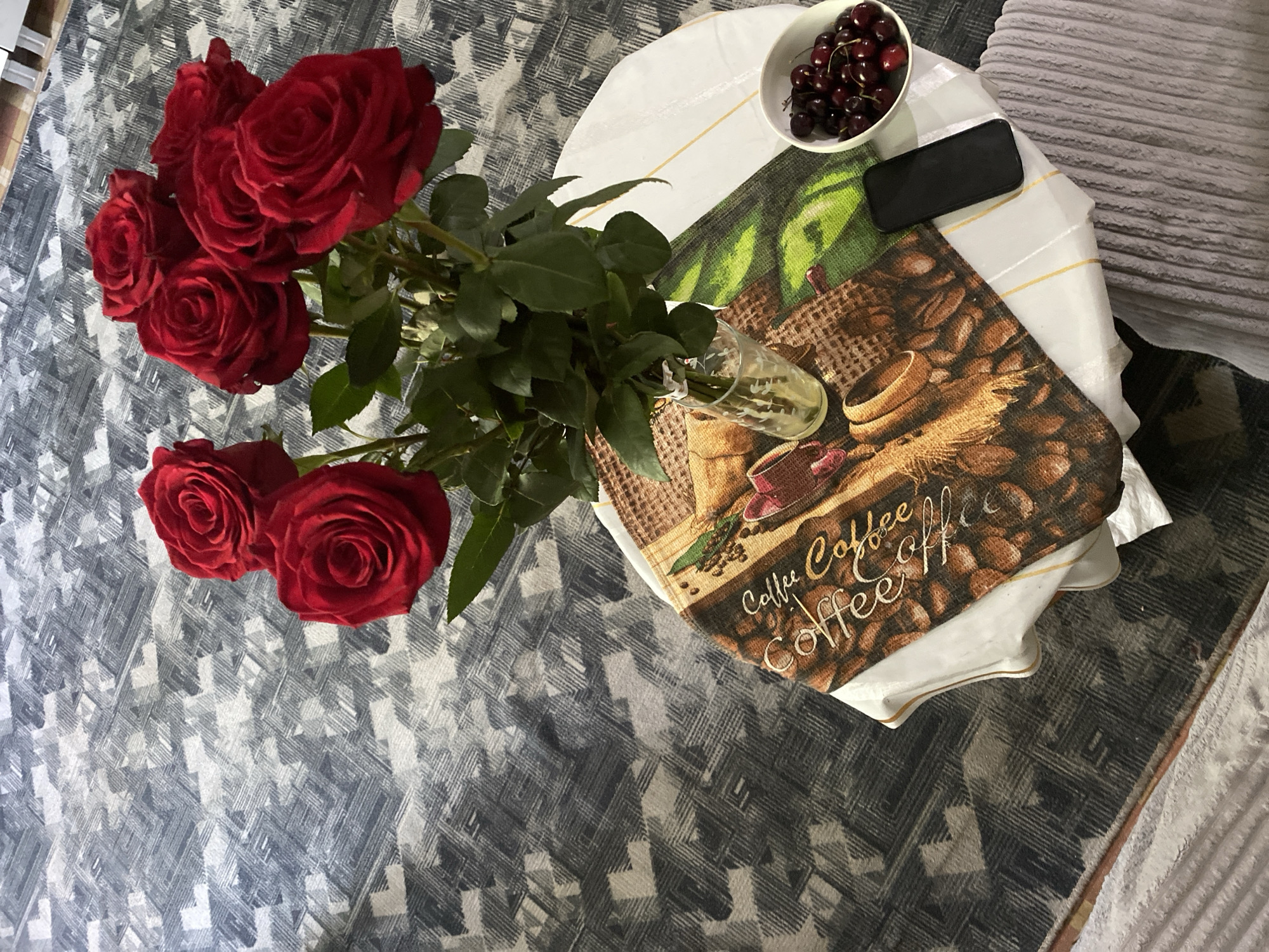 In Chernogorsk, Russia, a still life photograph captures a bouquet of vibrant red roses arranged in a glass vase. The vase rests on a table covered with a tablecloth adorned with a coffee-themed print, featuring coffee beans and cups. Beside the vase, a small white bowl is filled with dark red cherries. A black smartphone lies on the tablecloth, adjacent to the bowl. The table itself is set on a patterned rug. The scene is illuminated by ambient lighting, casting subtle shadows. There are no individuals present in the photograph.