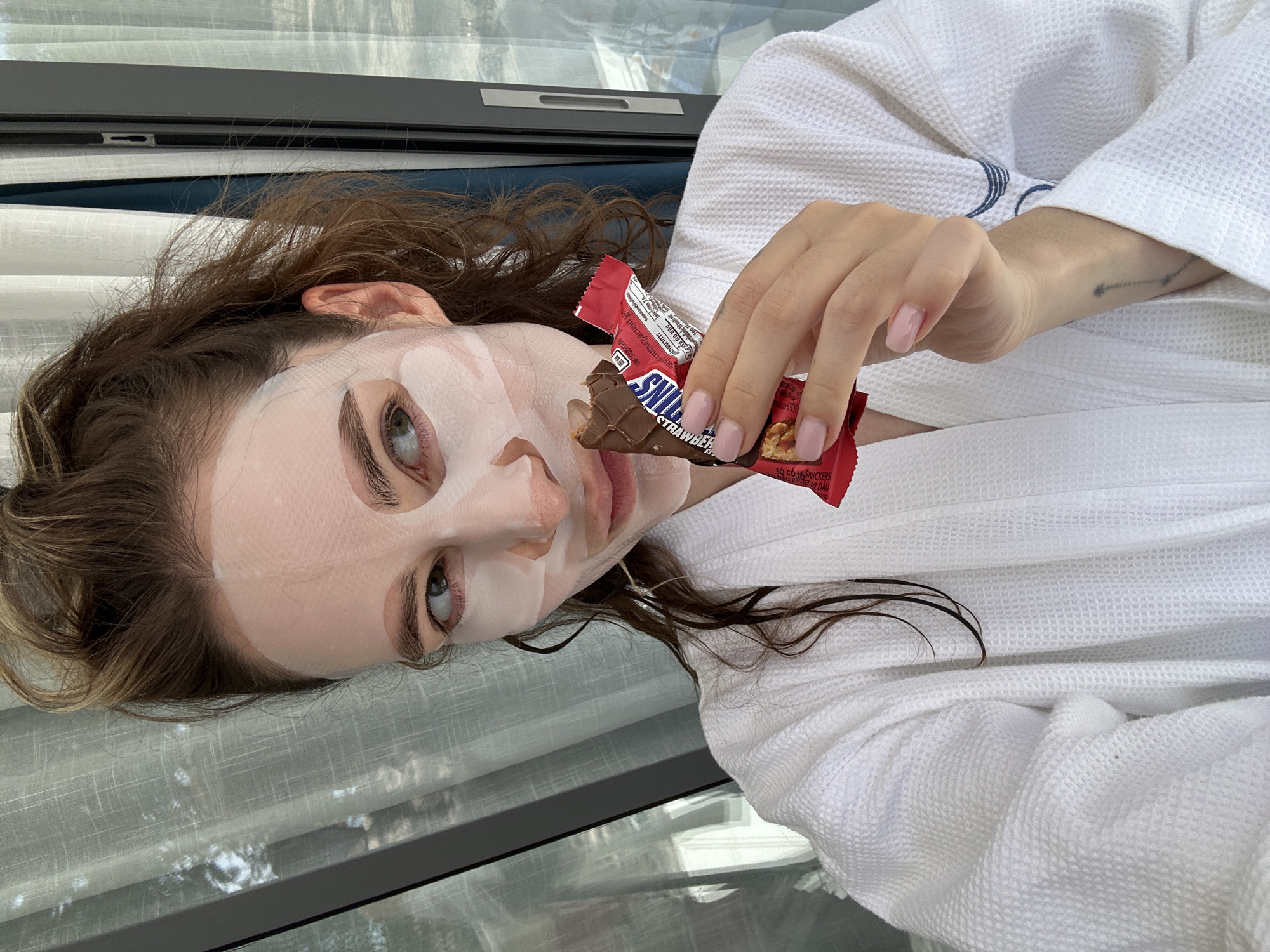A female individual is depicted in an indoor setting, likely a room with a large window in Choeng Thale, Thailand. She has light brown, wavy hair, some strands appearing wet, and light-colored eyes. Her face is covered by a white sheet face mask, leaving her eyes, nostrils, and mouth area exposed. She is wearing a white waffle-weave bathrobe with dark blue embroidered detailing visible on the right arm. In her right hand, she holds a partially consumed Snickers chocolate bar, identifiable by its red and blue wrapper and the exposed chocolate, caramel, and peanuts. Her fingernails are painted light pink. A faint tattoo is visible on her left forearm. She is looking towards her left. The background shows a large window frame, with sheer white curtains covering the view beyond, and a dark blue curtain section on the right.
