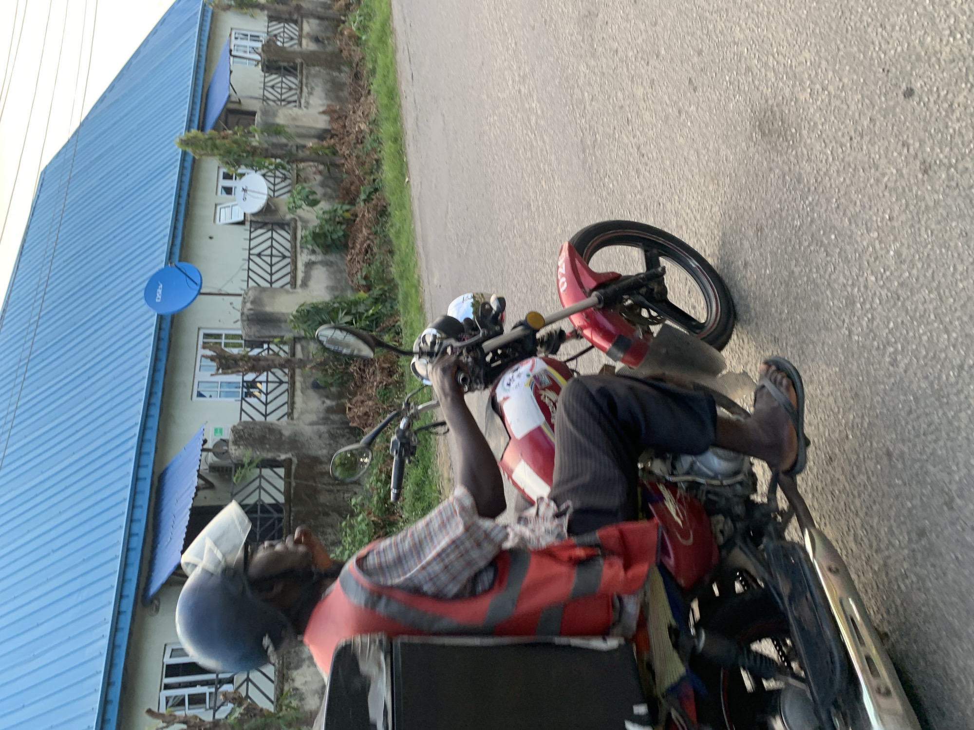 A man wearing a helmet and an orange safety vest is riding a red and white motorcycle on a paved road in what appears to be Abuja, Nigeria. The man is wearing sandals. The motorcycle has a delivery box attached to the back. Buildings with blue roofs and satellite dishes are visible in the background. The road is asphalt and slightly cracked. Green foliage is present alongside the buildings.