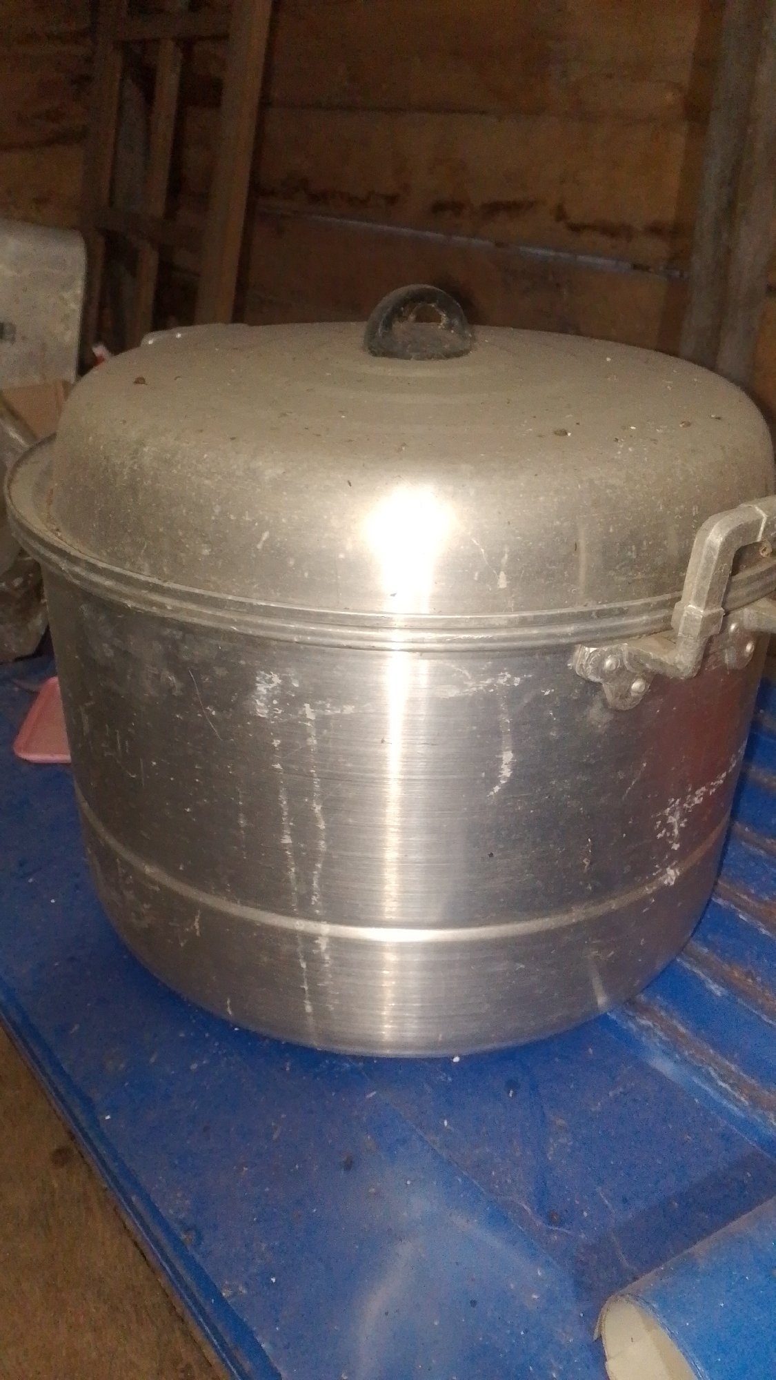 The image shows a large, silver, metal steamer pot with a black knob on top of its lid. The pot is sitting on a blue surface, which appears to be a table or a shelf, in what looks like a rustic or storage area. The background consists of wooden planks, suggesting an indoor setting with unfinished construction. There are no people visible in the image. The lighting is somewhat dim, with a bright reflection on the lid of the pot. The overall condition of the pot appears to be used, with some scuff marks and streaks on its surface. The environment seems to be a storage space, possibly a kitchen or a shed, in Olak-Olak Kubu, Indonesia, given the context.