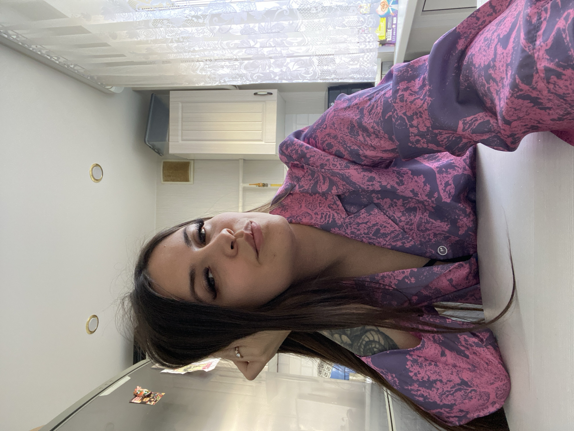 This media file depicts a young woman in a domestic indoor setting, identified as Chaikovsky, Russia. The image, which is rotated 90 degrees clockwise, shows the woman lying in the foreground, looking directly at the camera with a neutral and calm expression. She has long, dark brown hair and prominent eyelashes. She is wearing a purple and pink patterned, collared top, possibly a pajama set or a casual blouse. A ring is visible on her left ring finger, and part of a dark tattoo is visible on her left arm or shoulder.
The background suggests a modern kitchen or an open-plan living space. White kitchen cabinets are visible, along with a window covered by white patterned blinds. To the left, a portion of a refrigerator is seen, with a small, colorful decorative item or magnet attached to its door. A small, golden-rimmed circular light fixture is mounted on the white wall. In the distance, behind the woman, shelves hold a few indistinct items, including some product packaging with blurry, illegible text, and a yellow cylindrical object.
The lighting indicates it is daytime, with ambient light filtering through the window. The overall scene conveys a sense of relaxed domesticity. No clear, legible text is visible within the image beyond the faint, unidentifiable text on the packaging.