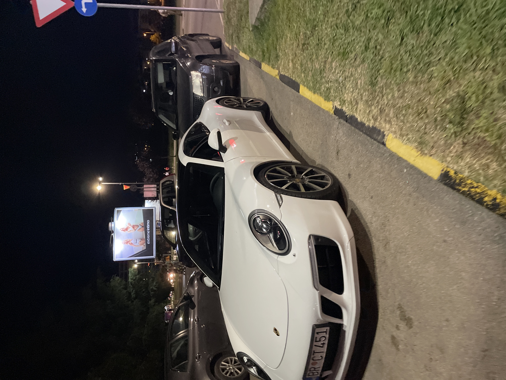 At night, on a paved road in Bar, Montenegro, a white Porsche 911 is parked, facing right. The car's license plate reads "BR CT 451". To its right, a dark-colored SUV is also parked. In the background, a billboard displays an advertisement for "extreme Intimo" featuring two women in swimwear. Traffic lights and streetlights illuminate the scene, with trees and buildings visible in the distance. A pedestrian crossing sign is positioned on the right side of the image. The ground is paved with asphalt, bordered by a grassy area with a yellow and black curb.