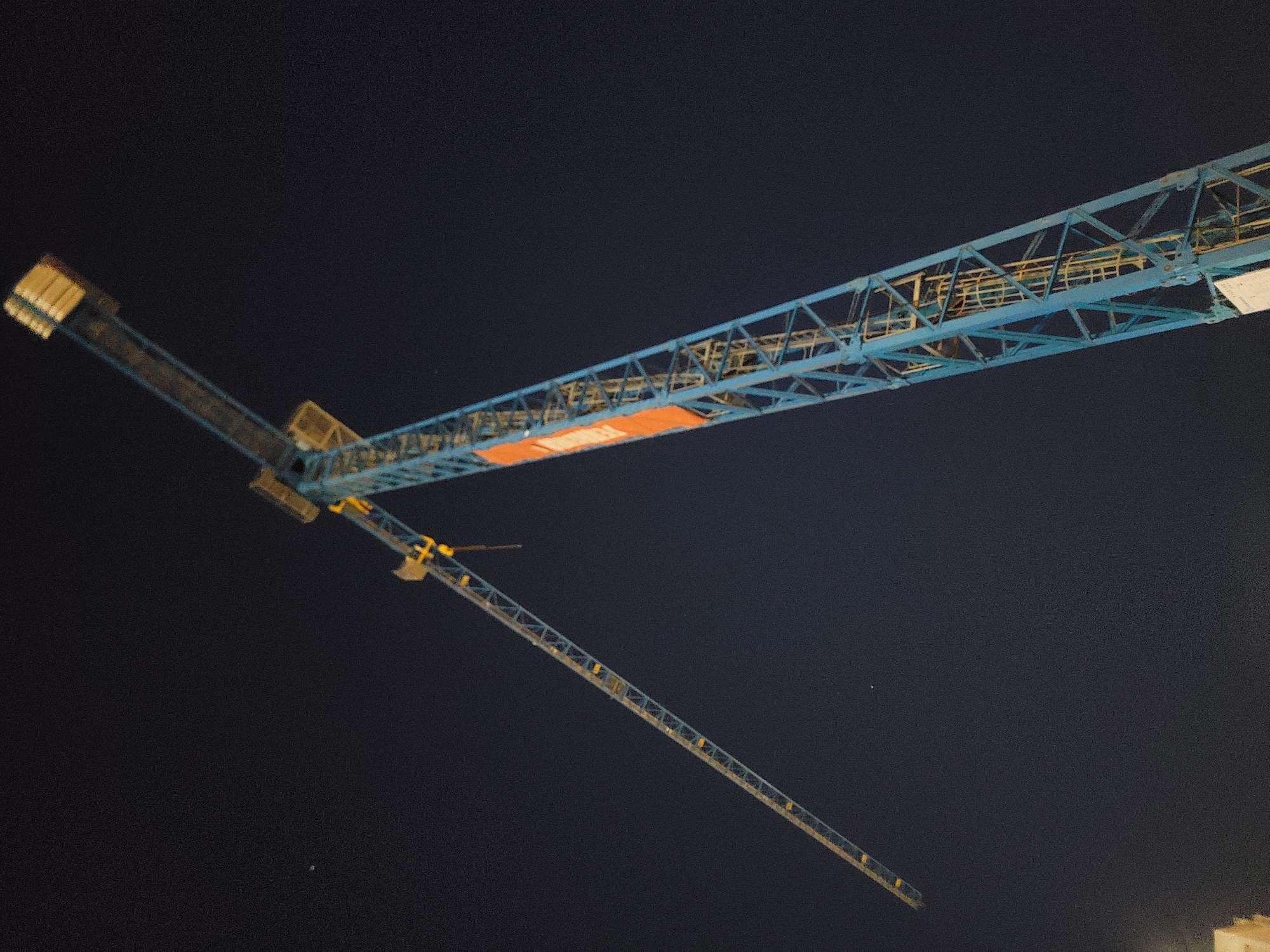 The image shows a close-up, low-angle shot of a blue construction crane against a dark night sky. The crane's structure is visible, with its long jib extending diagonally upwards to the left and its main tower rising vertically on the right. Yellow accents are present on the crane's structure and at the top of the jib. An orange panel with white text is affixed to the main tower. A faint, white speck, possibly a distant light or star, is visible in the sky to the left of the jib. The bottom left of the image shows a portion of a building with light-colored walls. The scene is set in Quarteira, Portugal, likely at a construction site.
