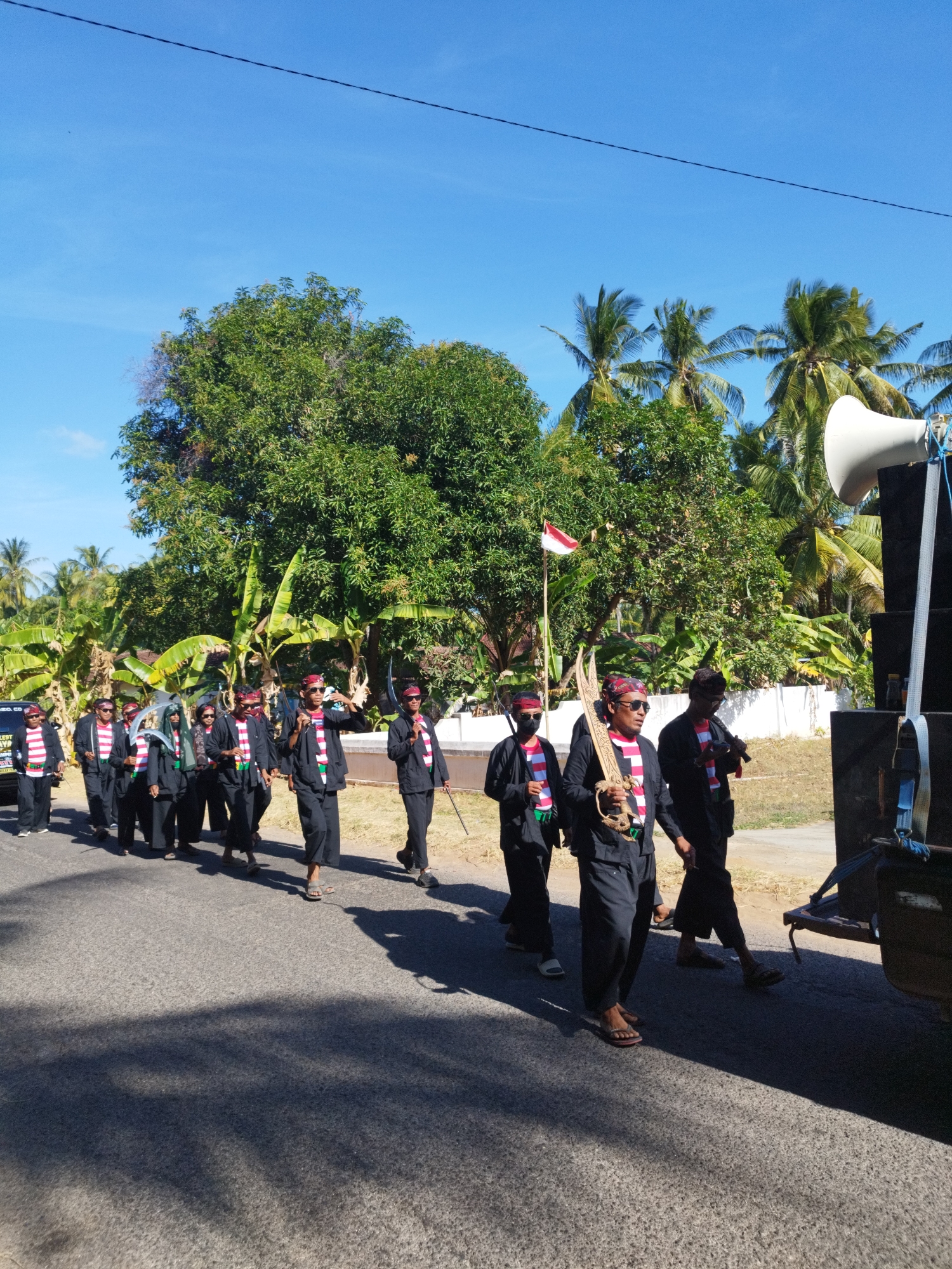 A group of men, dressed in traditional black clothing with red and white striped sashes and red headbands, are walking in a procession on a paved road in Andulang, Indonesia. The men are carrying what appear to be ceremonial weapons, possibly swords or daggers, and some are wearing sunglasses. The procession is accompanied by a large speaker system mounted on a trailer. In the background, there are trees, including palm trees, and a clear blue sky. The sun is casting long shadows on the road, indicating it is likely daytime. A small Indonesian flag is also visible on a pole behind the group. The overall scene suggests a cultural or ceremonial event taking place outdoors.