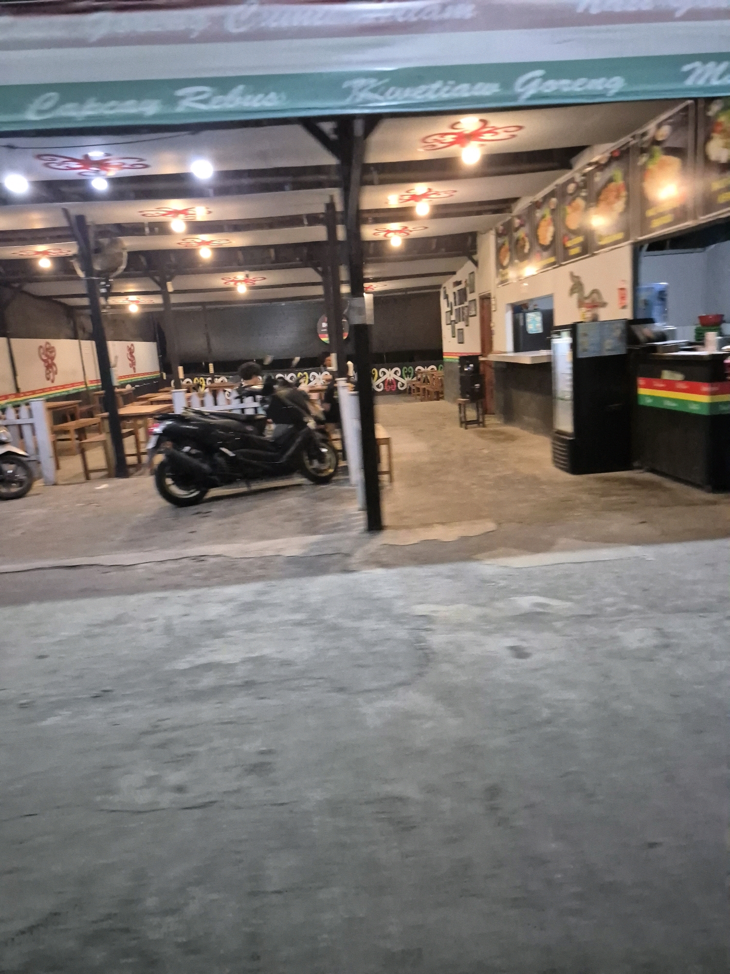 The image shows the exterior of a restaurant in Pabelan, Indonesia, at night. A green awning with white lettering reads "Capcay Rebus" and "Kwetiau Goreng". Inside, tables and chairs are visible, and a motorcycle is parked in front of the establishment. The restaurant appears to be open, with lights illuminating the interior. The overall atmosphere is casual and typical of a street-side eatery.