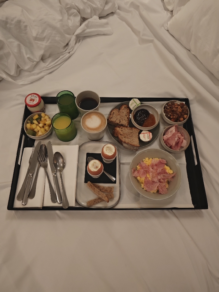 A black tray laden with a breakfast feast rests on a white bed. There's a mix of beverages: juice, coffee, and a latte. Food items include fruit salad, yogurt, toast, jam, butter, sliced bread, hard-boiled eggs, scrambled eggs topped with ham, a dish of nuts, and sliced meat. Cutlery is neatly arranged on a napkin. The bedding is rumpled, suggesting a cozy and inviting setting.
