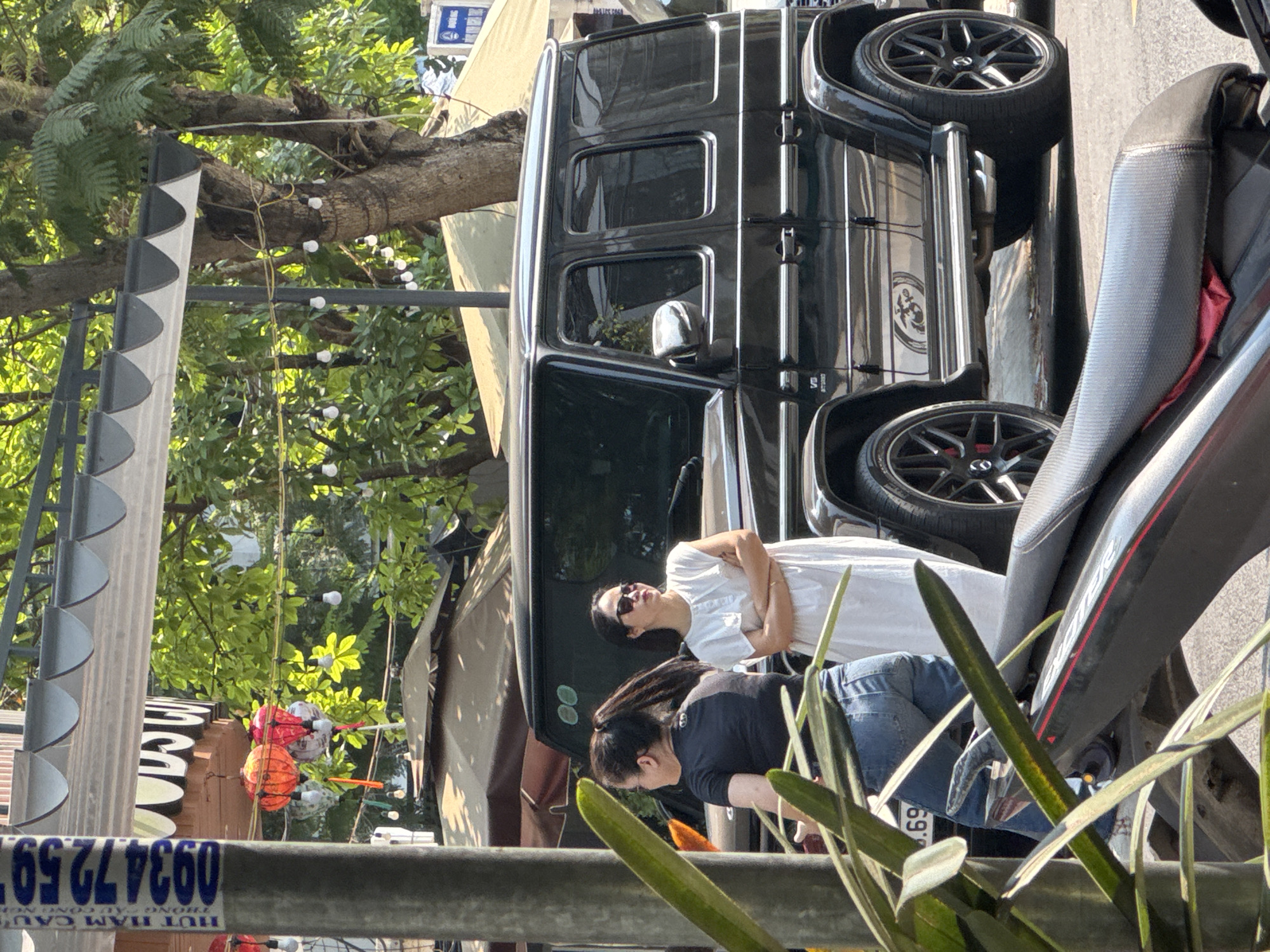 The scene captures a moment on a street in Đà Nẵng, Vietnam. A black Mercedes-Benz G-Wagon is parked on the side of the road, positioned at an angle to the frame. Two women are standing near the vehicle; one is wearing a white dress and sunglasses, her arms crossed. The second woman is wearing a dark t-shirt and blue jeans, bending over. To the left of the frame is a vertical pole with affixed signage bearing the phone number "0934 72 59 60" and Vietnamese text. Above this pole is a partial view of a restaurant or shop façade. Lanterns are hung up for decoration. Dense foliage from trees is present, filtering the sunlight. A parked motorbike is positioned towards the right side of the frame.