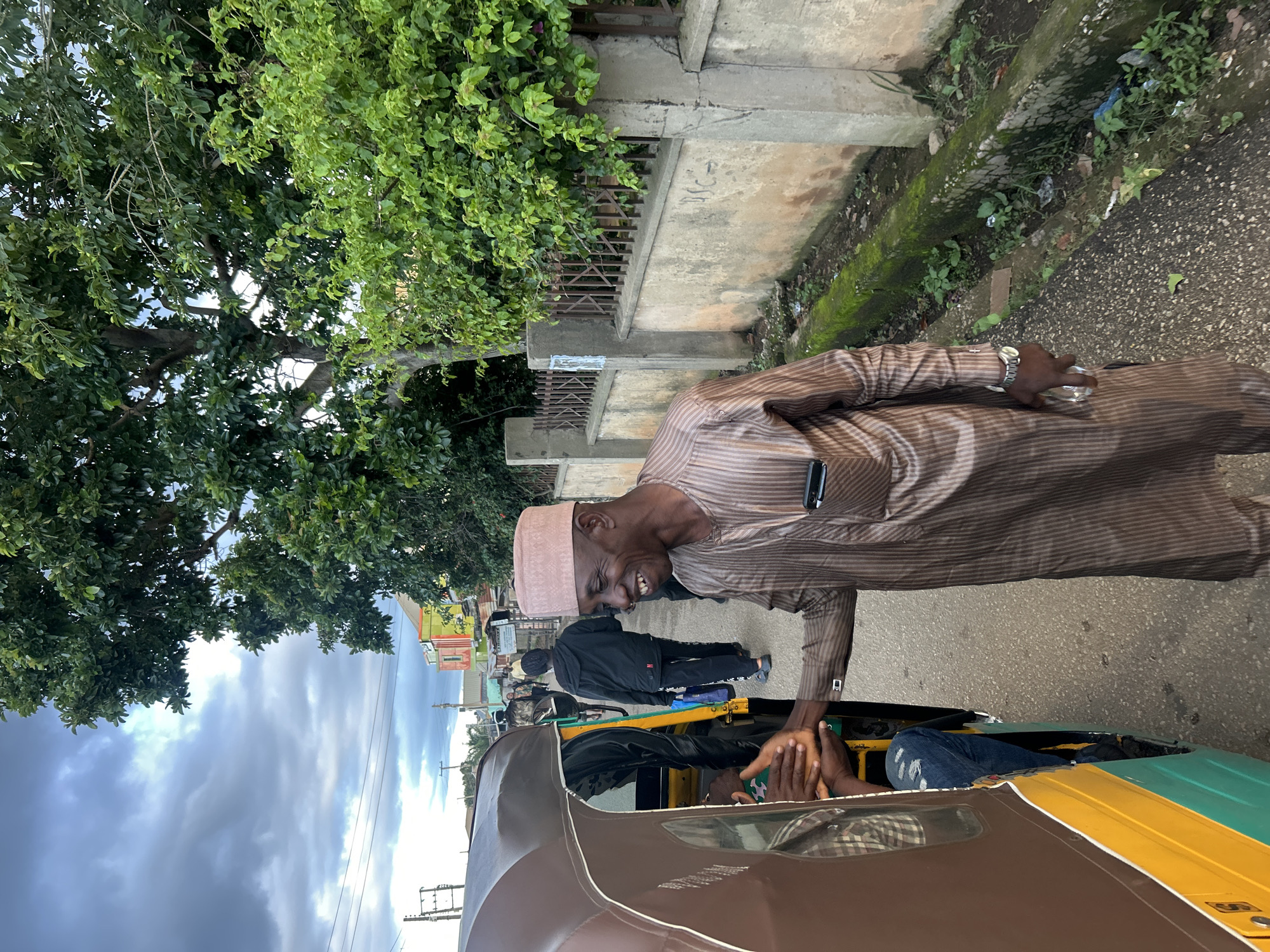 A man in a brown pinstripe kaftan and a matching cap is shaking hands with someone inside a three-wheeled auto-rickshaw. He is smiling. The auto-rickshaw is brown and yellow. The man is standing on a paved road in front of a concrete wall with a metal fence on top. There is a drainage ditch beside the wall filled with moss and weeds. In the background, there are buildings, trees, and a cloudy sky. The scene appears to be in Jos, Nigeria.