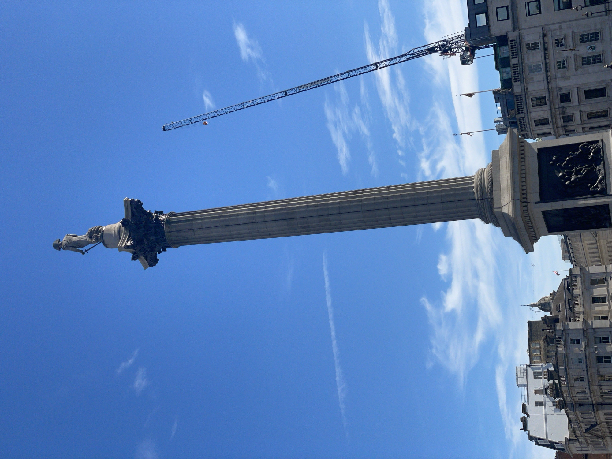 This media file captures a low-angle view of Nelson's Column in Trafalgar Square, London. The iconic monument, a tall granite column topped with a statue of Admiral Lord Nelson, stands against a bright blue sky dotted with wispy white clouds. The sun appears to be high in the sky, suggesting it is midday on a clear day.
To the right of the column, a tall, multi-story building with many windows is visible, indicating an urban setting. A construction crane extends across the upper portion of the frame, its metallic structure stark against the sky, suggesting ongoing development or maintenance in the area. The base of Nelson's Column features ornate reliefs, though the details are partially obscured by shadow. A small statue of a person is perched on top of the column, representing Admiral Nelson.
The scene is dominated by the imposing presence of the column and the vastness of the sky, creating a sense of scale and grandeur. The weather appears to be fair, with a few scattered clouds. The presence of buildings and the famous landmark firmly place the location as Trafalgar Square in London, United Kingdom. No visible text is present in the image.