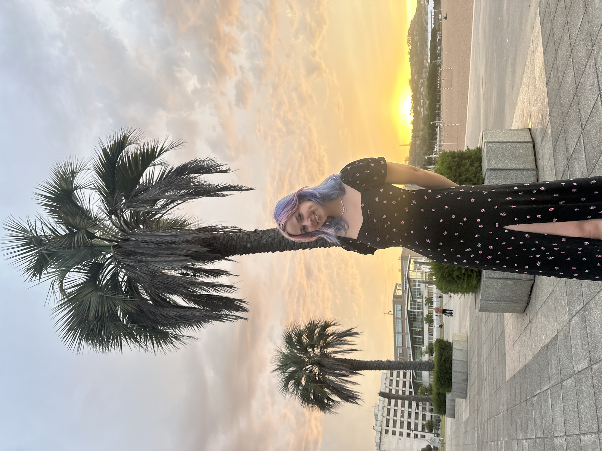 A young woman with dyed pink and blue hair stands in front of a sunset on a paved area near a beach, likely the Playa de San Lorenzo in Gijón, Spain. She is wearing a black dress with a floral pattern and a thigh-high slit, and a pearl necklace. Two palm trees are visible behind her, along with some modern buildings in the background. The sky is filled with dramatic clouds tinged with the orange and yellow hues of the setting sun. In the distance, a beach is visible with some people and what appear to be soccer goals.