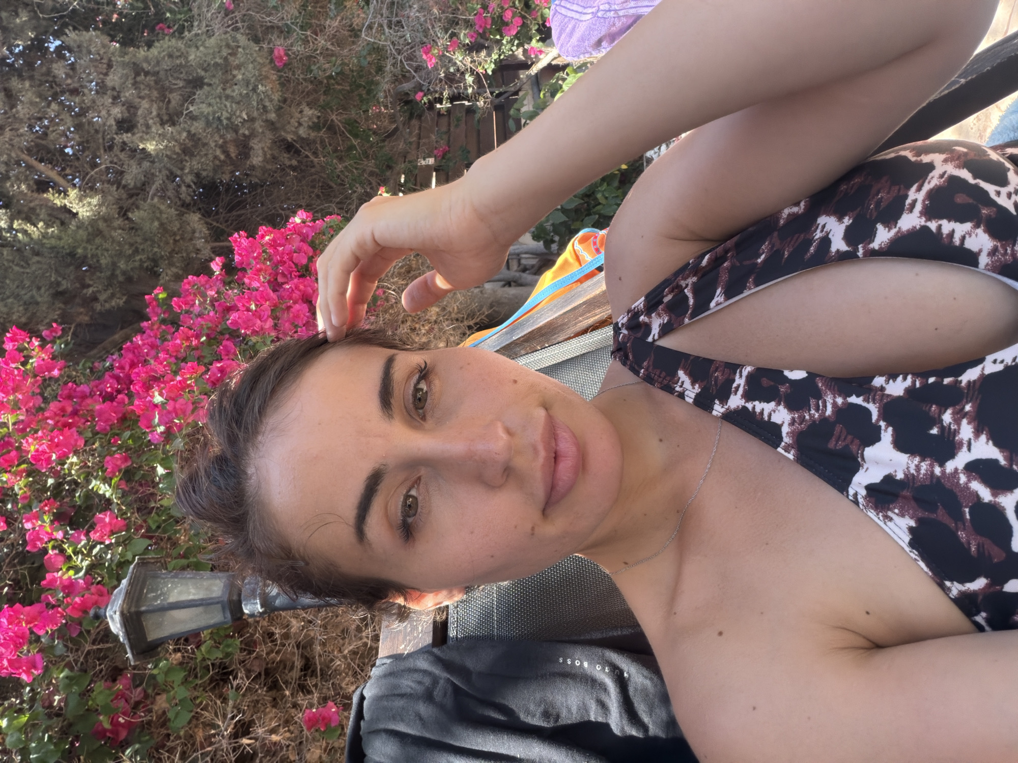 A woman is taking a selfie outdoors, possibly in Mazotos, Cyprus, given the presence of bougainvillea flowers and a light post. She is seated, wearing a leopard-print one-piece swimsuit with a cutout. Her hair is pulled up, and she is touching her forehead with her right hand. To her left, a dark hooded jacket is visible, and to her right, a towel and part of a beach chair are in the background. The overall impression is a relaxed, sunny day.