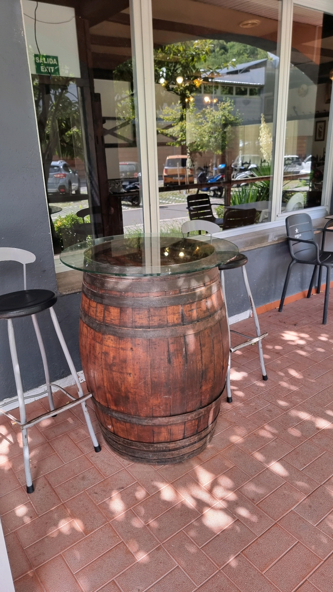 An outdoor seating area is depicted, featuring a table made from a wooden barrel topped with a circular glass surface. Two high stools, one white and one with a metal frame, are positioned around the table. In the background, through a large glass window, a street scene is visible, including parked cars, motorcycles, and buildings. A green "EXIT" sign is also visible in the window. The flooring is tiled with a reddish-brown pattern. The image appears to have been taken during the daytime, with dappled sunlight casting shadows on the ground.