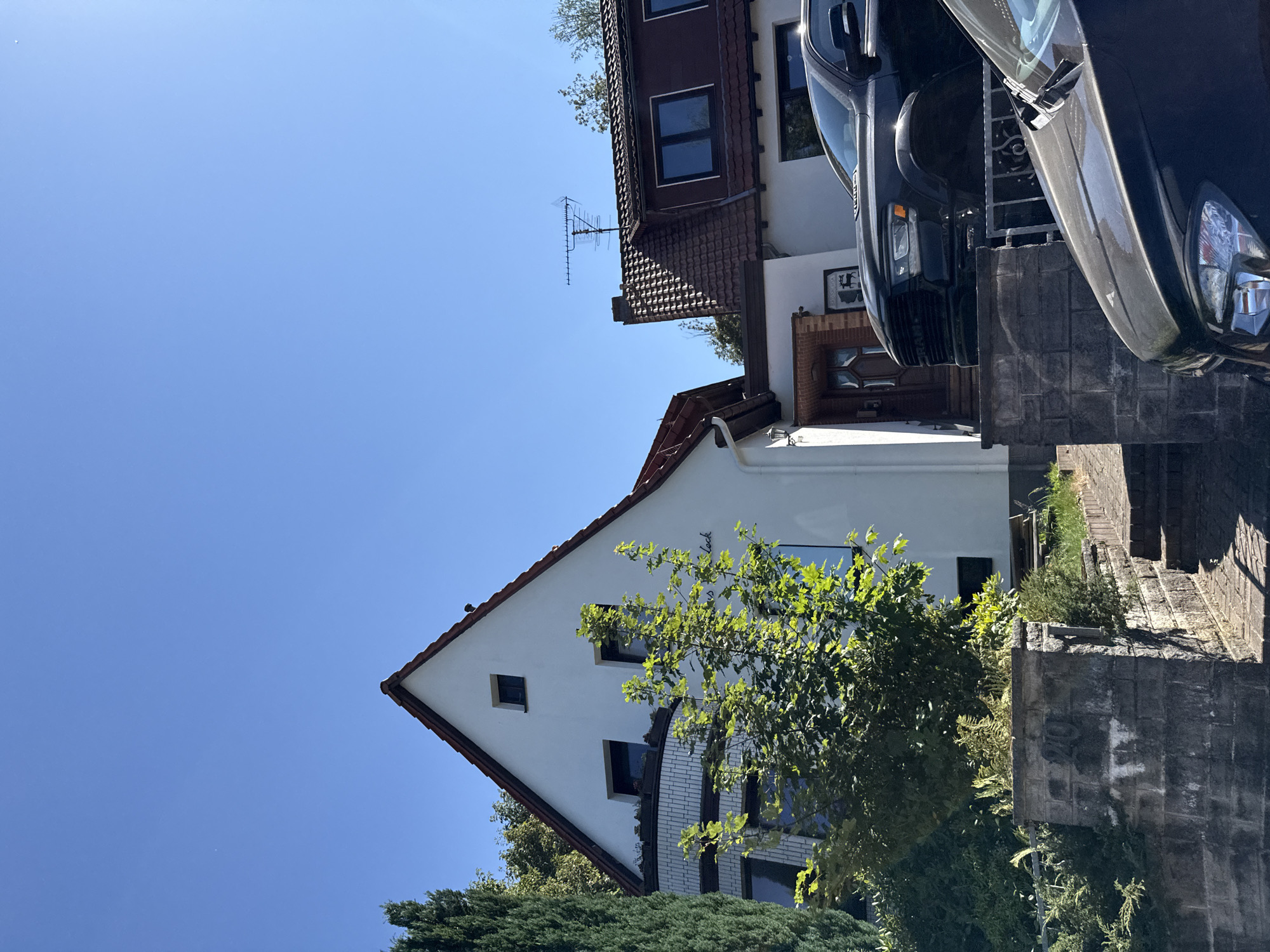 The image depicts a residential property in Bad Sachsa, Germany. A white, two-story house with a red-tiled roof is partially visible, along with an adjacent building with a similar roofline. A dark-colored truck and another dark-colored car are parked in the foreground. Green foliage and trees surround the buildings. The sky is clear and blue. A small sign with text is visible on the white house.