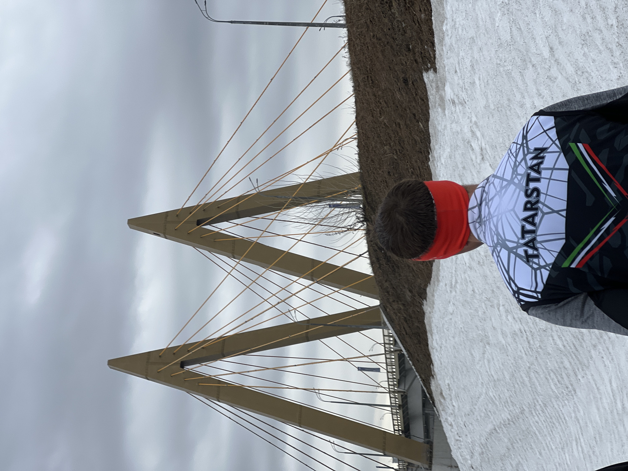 A low-angle, medium shot depicts a person wearing a red headband and a white, grey, black, and green sports shirt with the word "TATARSTAN" written on it, positioned next to what appears to be a snow-covered ground or a building with a snow-like texture on the side. In the background, there is a cable-stayed bridge with two prominent A-shaped towers and numerous cables extending from the towers. The sky is overcast. The bridge is most likely the Millennium Bridge in Kazan, Russia. A streetlight pole is visible at the top of the frame.