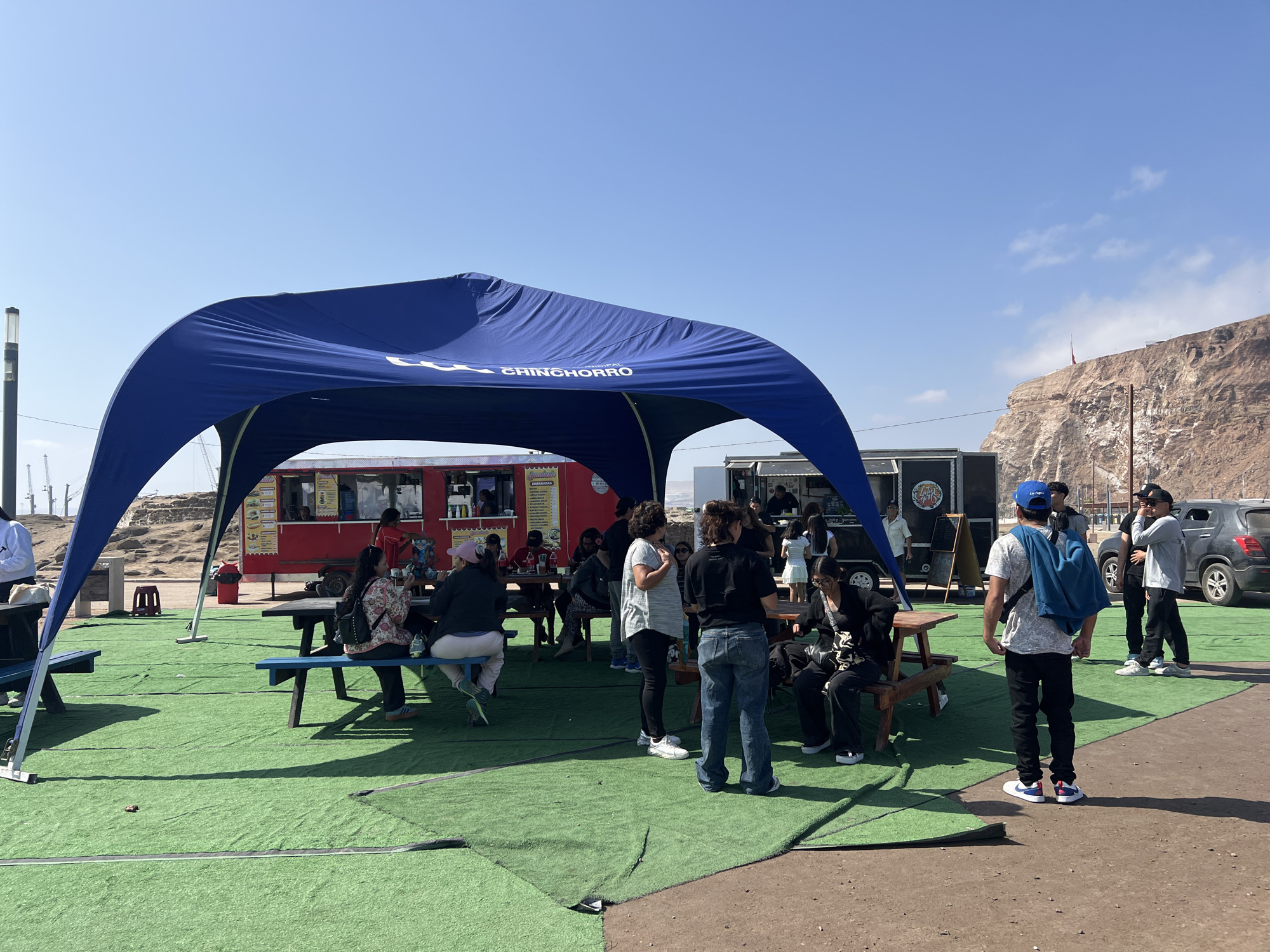 This image captures a scene in Arica, Chile, likely at a recreational area or food court. In the foreground, a group of people are gathered under a large blue canopy. The canopy has text on it that reads "CHINCHORRO". Several individuals are seated at picnic tables, which are placed on artificial green turf. Other people are standing and conversing.
Behind the canopy, there are several food trucks. One food truck is red and appears to be selling empanadas, with signage in Spanish visible. Another food truck is black with white lettering. In the background, a large, rocky cliff face dominates the landscape, a characteristic feature of Arica. The sky is clear and blue. A dark-colored SUV is parked to the right of the scene. The ground is a mix of dirt and the artificial turf. The overall impression is of an outdoor gathering with food vendors, set against a natural, arid backdrop.