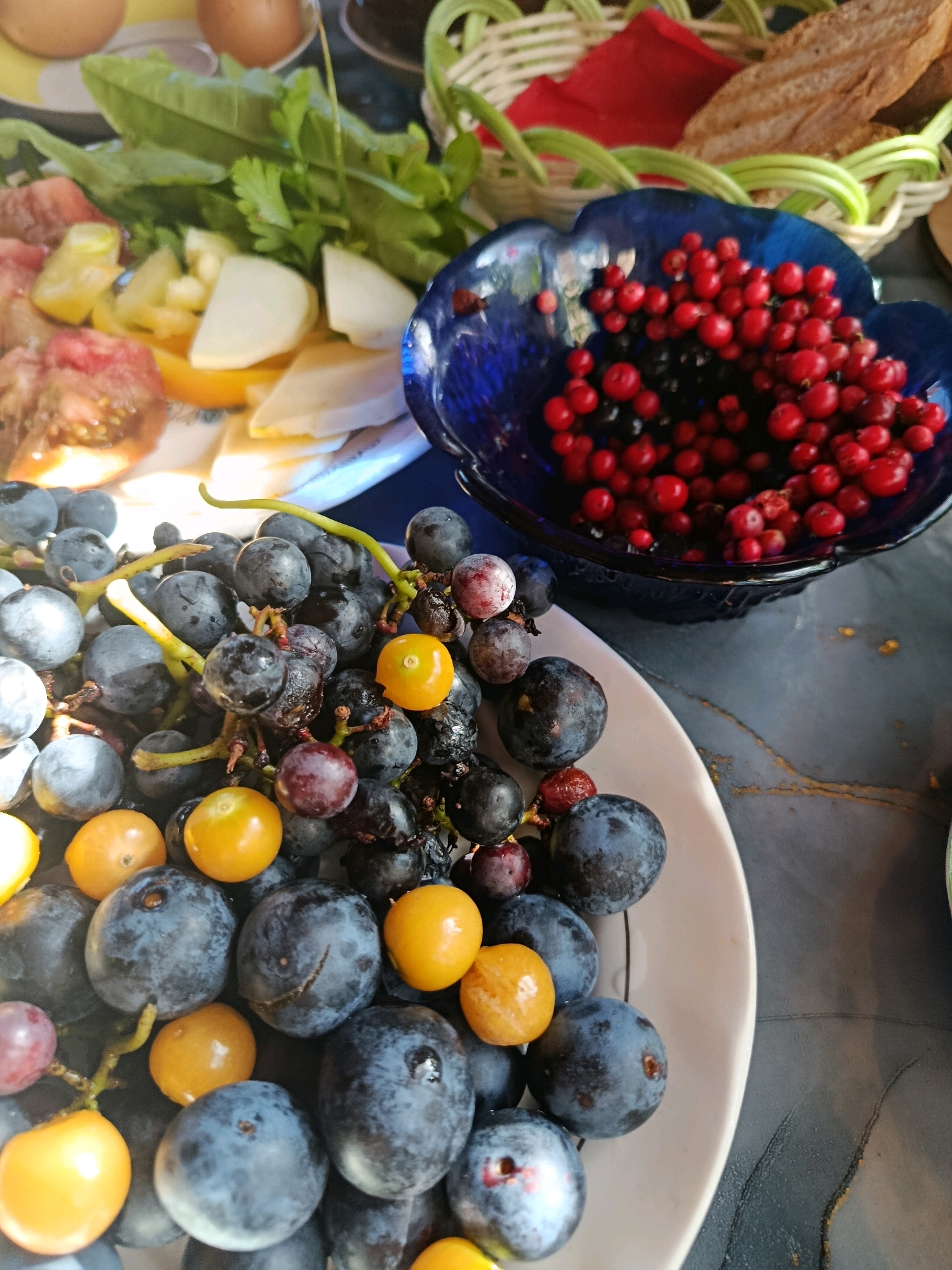 A platter of grapes and other fruits, along with a basket of bread and red cloth, are arranged on a dark, textured surface. The grapes are a mix of dark blue and purple, with some yellow and red fruits interspersed. A blue bowl filled with red berries and some dark berries is positioned to the right of the platter. In the background, a basket with woven sides contains slices of bread and a red cloth. Some green leafy vegetables and sliced tomatoes are also visible on the platter. The lighting suggests an indoor setting, possibly a dining table.